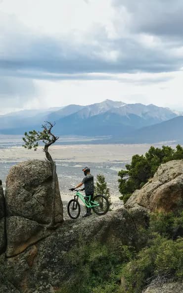 A person wearing a helmet and standing with a bicycle on a large rock formation overlooking a vast landscape of mountains and valleys. The sky is cloudy, and there is a small tree growing out of the rock nearby.