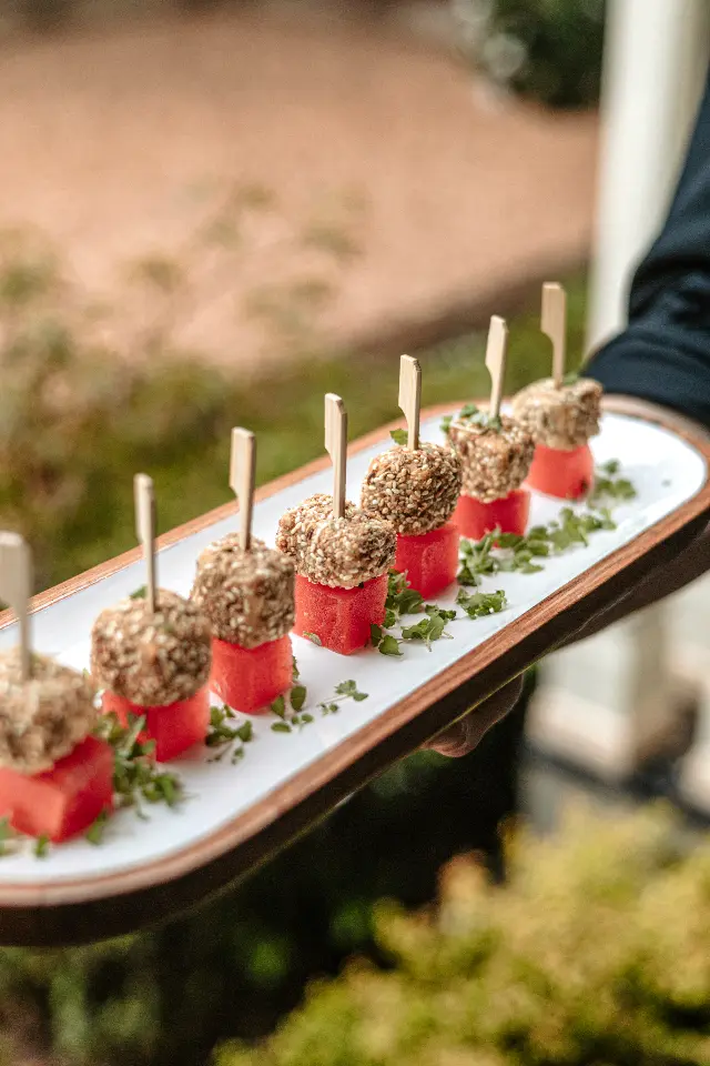 A rectangular platter holds a row of appetizers, each consisting of a cube of watermelon topped with a sesame-coated ball, held together by a toothpick. The platter is garnished with small green herbs and is being carried by a person in dark clothing.