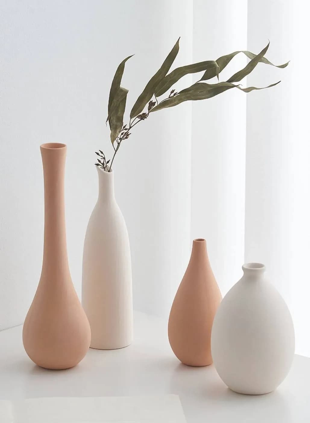 A minimalist arrangement of four ceramic vases in neutral tones of beige and white. Two tall, slender vases with leaves are in the back, while two shorter, round vases are in the front, all set against a soft, white backdrop.