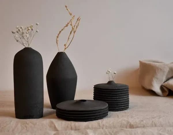 Two tall, black ceramic vases with minimalist dried flowers stand beside a stack of round, black ceramic plates with smaller vases on top. The setup is on a textured surface with a neutral-colored fabric in the background.