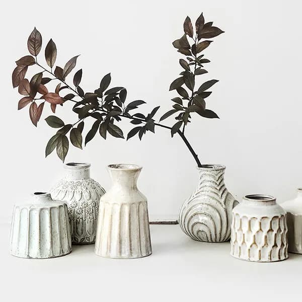 A minimalist display of six cream-colored ceramic vases with varying textures and designs. A branch with dark leaves is arranged among the vases, adding a touch of natural elegance against the plain background.