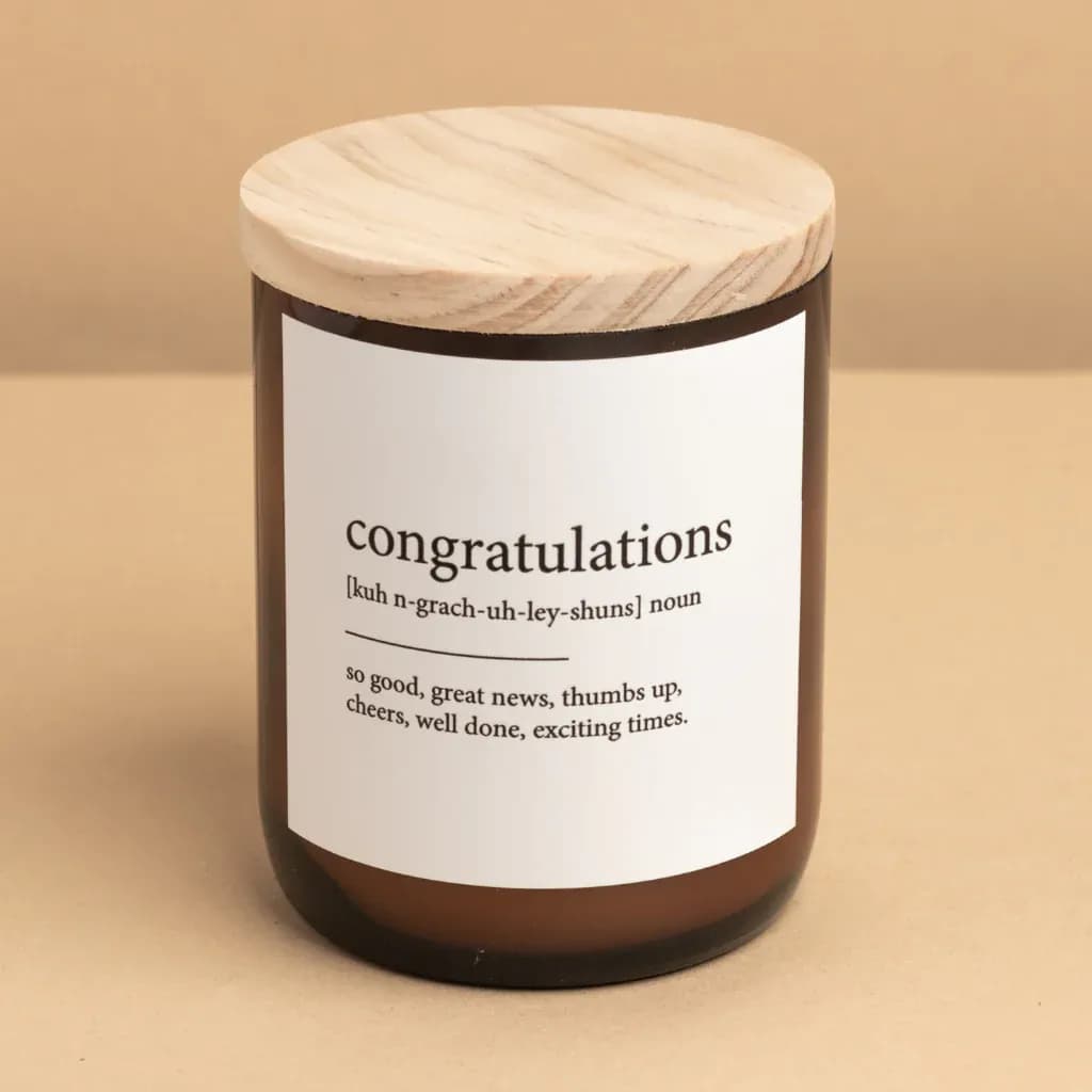 A brown jar candle with a wooden lid sits on a beige surface. The label on the jar reads "congratulations [kuh n-grach-uh-ley-shuns] noun - so good, great news, thumbs up, cheers, well done, exciting times.