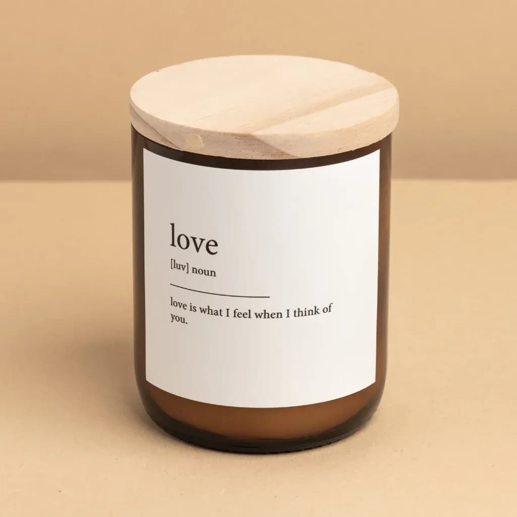 A brown candle with a wooden lid sits on a neutral background. The label on the candle reads: "love [luv] noun love is what I feel when I think of you.