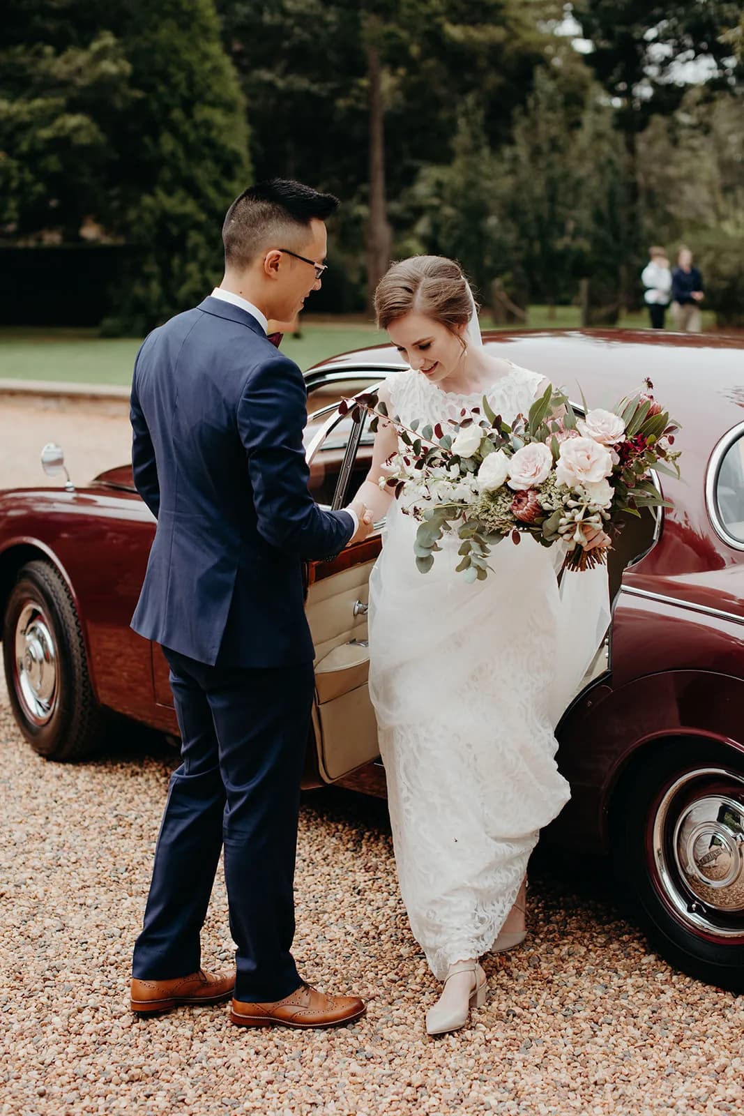 A bride in a white dress and groom in a blue suit stand next to a vintage maroon car. The groom holds the bride's bouquet of flowers as she steps out of the car. Trees and greenery are blurred in the background.