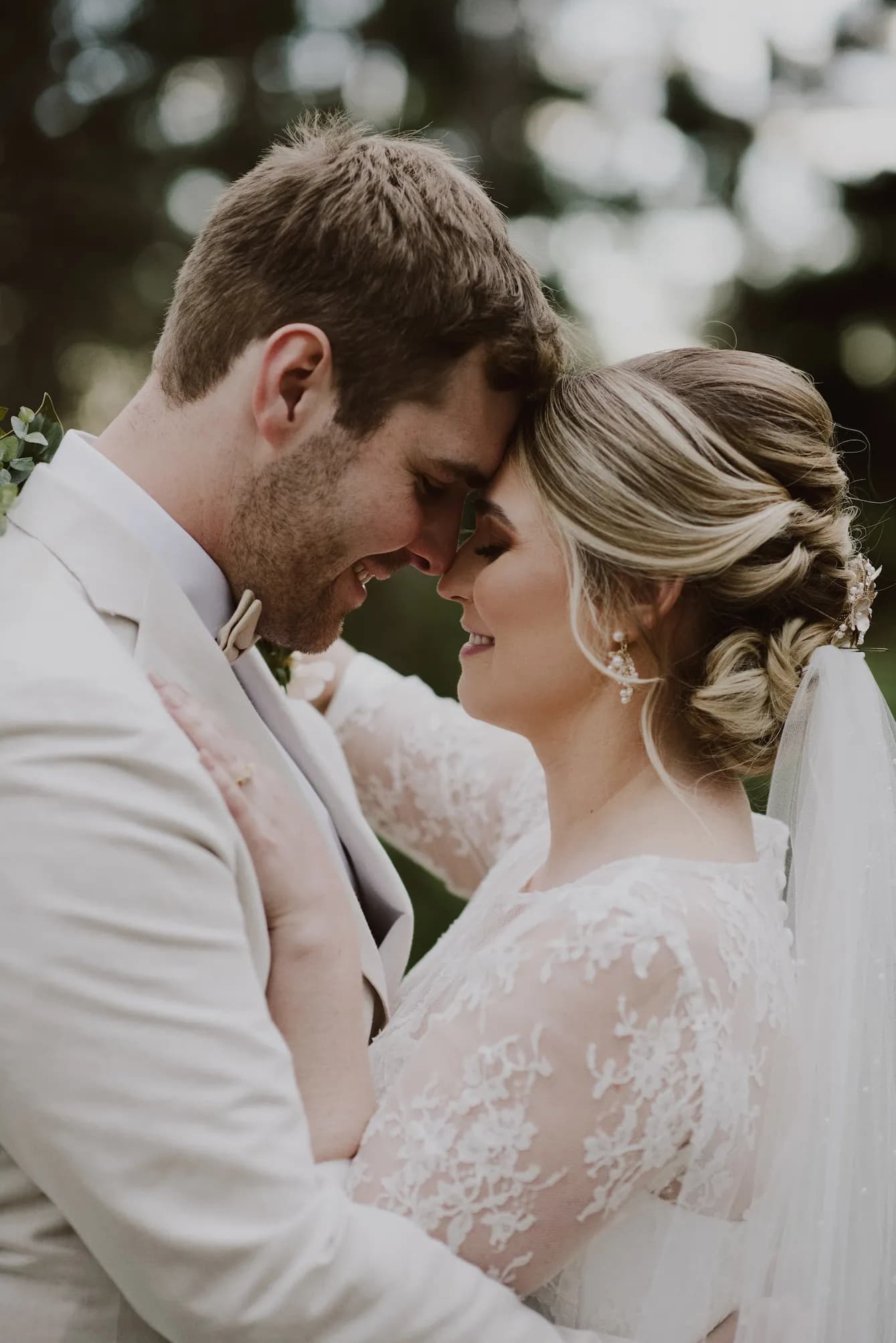 A bride and groom, dressed in wedding attire, stand closely with their foreheads touching and eyes closed, smiling. The bride wears a lace dress with a veil, and the groom wears a light-colored suit with a bow tie. They are surrounded by greenery in the background.