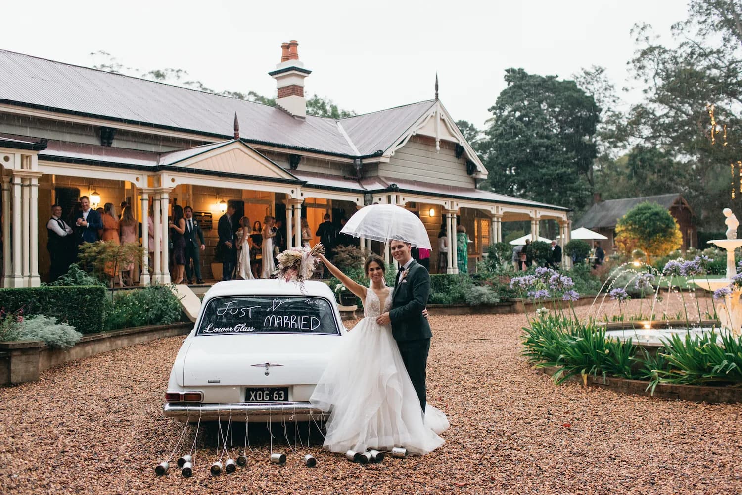 A newlywed couple stands next to a white car with "Just Married" written on the back, holding a transparent umbrella. They are in front of a large house with guests watching from the porch. The car has tin cans attached to the bumper. The area is decorated with flowers.