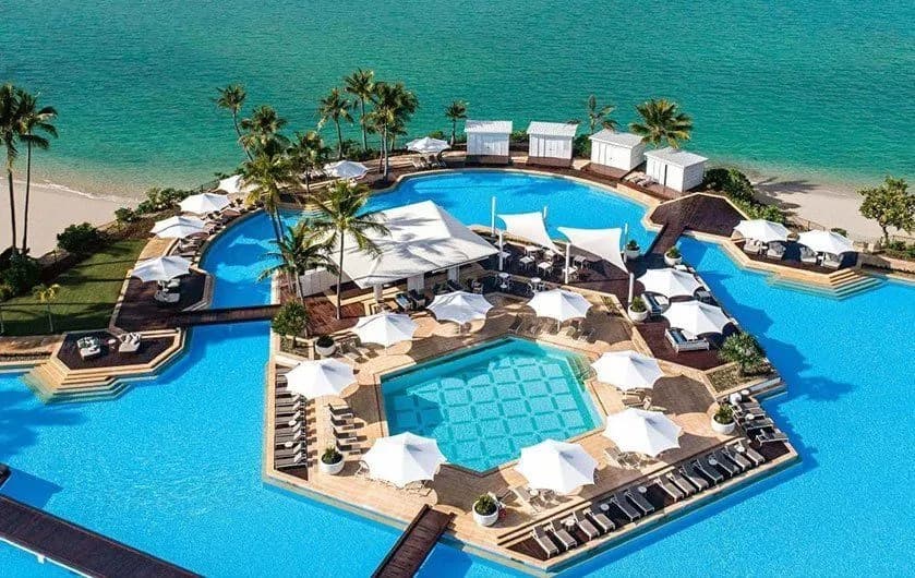 an aerial view of a large communal pool next to the ocean surrounded by sun loungers and palm trees