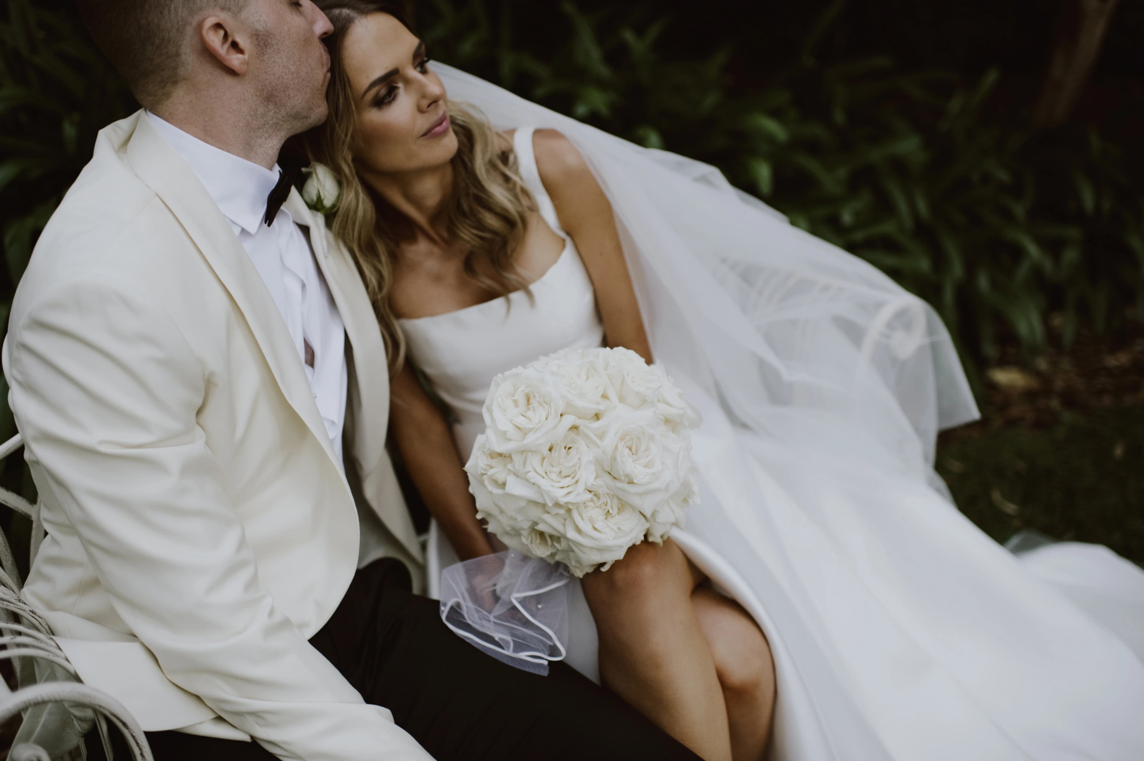 couple pose on a garden chair, bride stares into the sunset whilst holding bouquet of white roses