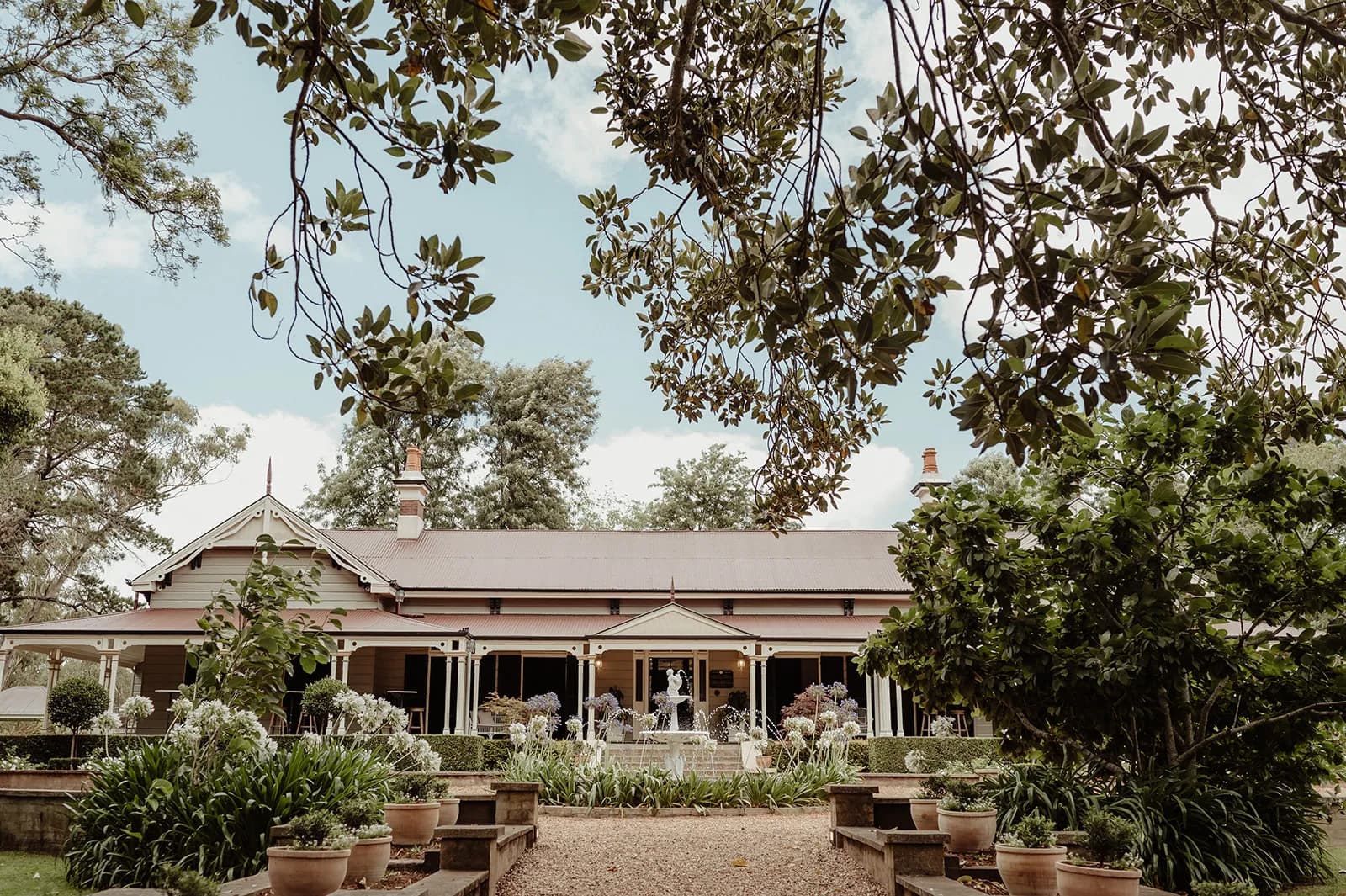 A charming, single-story house with a wide veranda is surrounded by lush greenery and tall trees. Potted plants and neatly trimmed bushes line the pathway leading to the entrance, creating a welcoming and serene atmosphere.