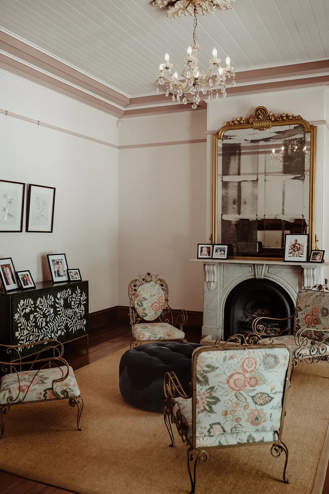 A cozy and elegant living room with ornate floral armchairs, a large black ottoman, a decorative black cabinet with framed photos and drawings, a chandelier, a large gold-framed mirror above a fireplace, and soft lighting from the chandelier.