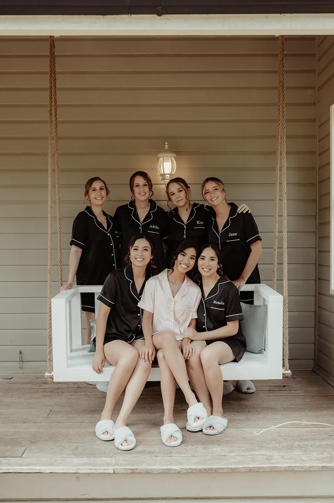 Six women are sitting on a porch swing. Five of them wear matching black pajamas with white trim, while the woman in the middle wears pink pajamas. They are all smiling and appear to be enjoying each other's company. The porch swing is suspended by ropes.