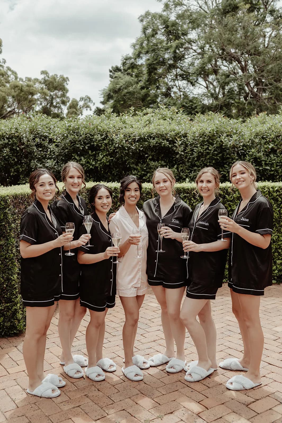 A group of seven women are standing outside on a brick patio, surrounded by greenery. Six of them are wearing matching black pajamas, while one in the center wears light-colored pajamas. They are all holding champagne glasses and smiling at the camera.