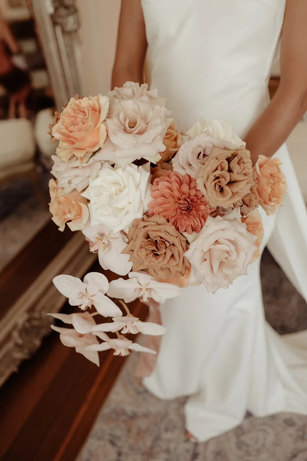 A bride holding a bouquet of flowers featuring shades of white, peach, beige, and pink. The bouquet has a mix of roses, dahlias, and orchids. The bride is wearing a white wedding dress, and part of a decorative mirror and a chair are visible in the background.