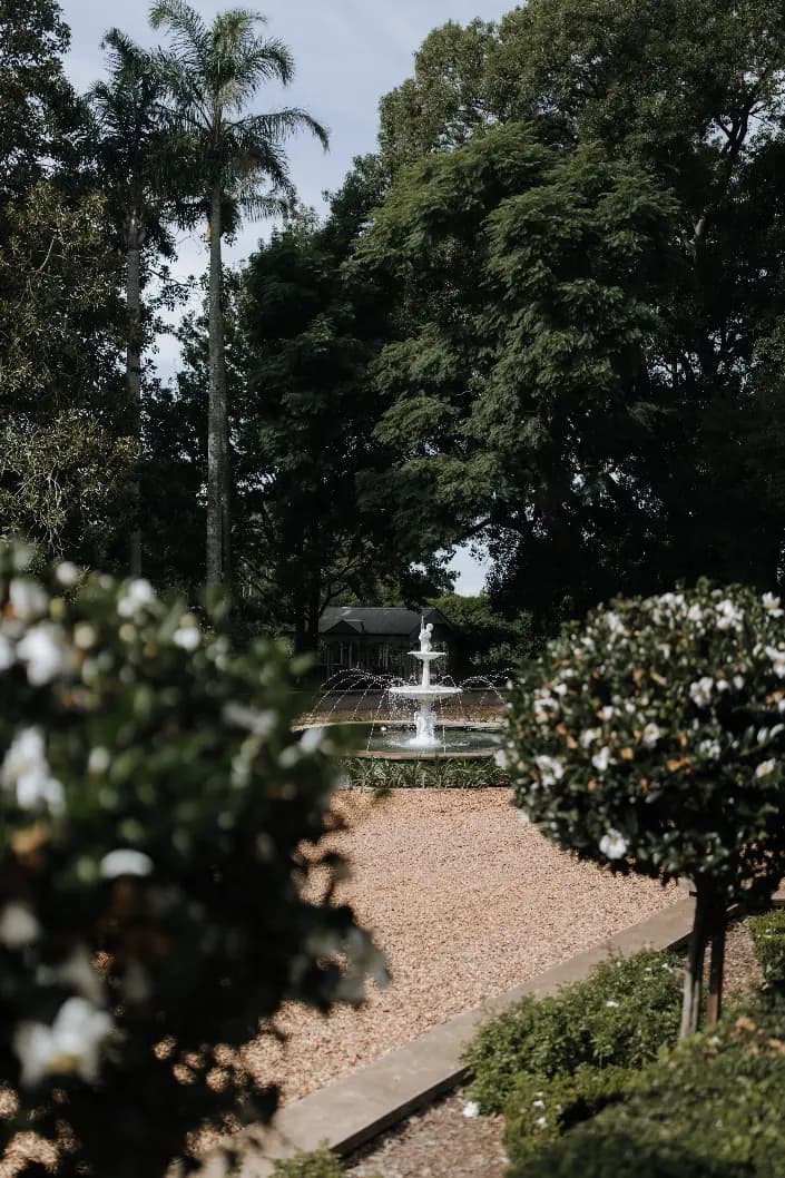 A white fountain stands at the center of a well-maintained garden surrounded by lush greenery. Palm trees and dense foliage are visible in the background, while neatly trimmed bushes and a gravel pathway frame the serene scene.