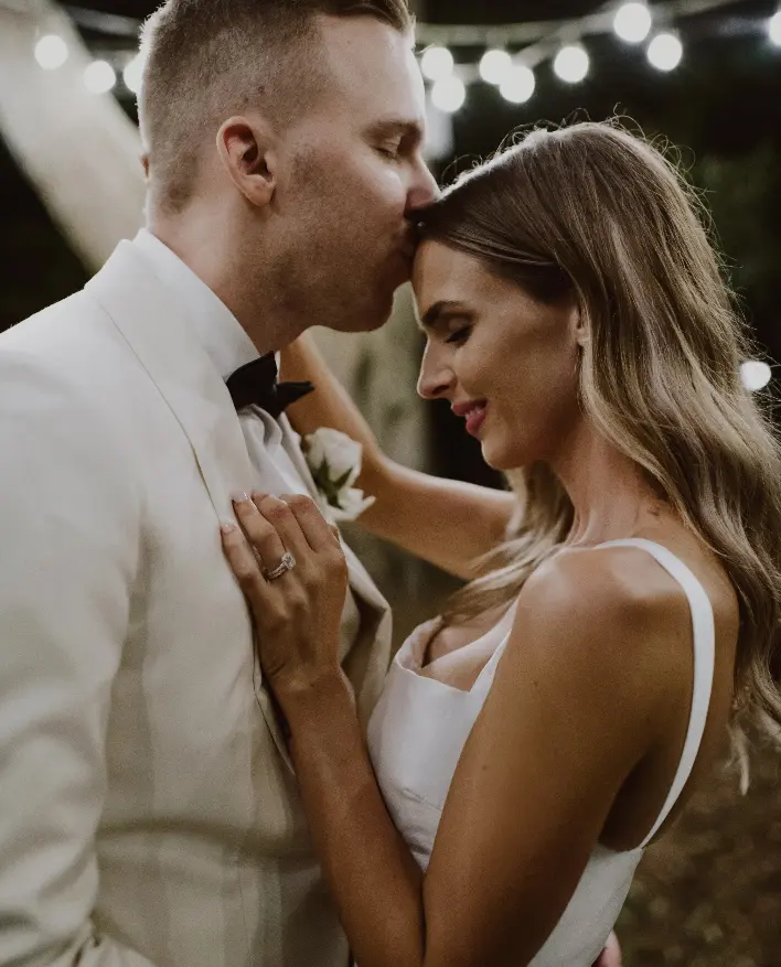 A groom in a white suit kisses a bride on the forehead during their wedding. The bride, with long wavy hair, smiles and closes her eyes while gently holding his lapel. String lights glow in the background, creating a romantic atmosphere.