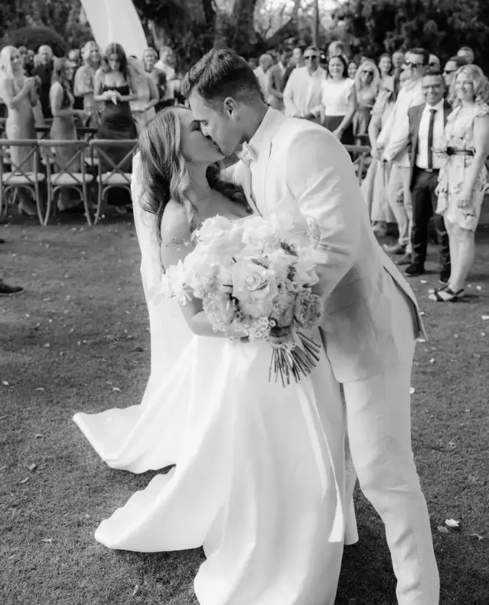 A newlywed couple share a kiss at their outdoor wedding ceremony. The bride holds a large bouquet of flowers, and guests stand in the background, watching and smiling. Both the bride and groom are dressed in white. The moment is captured in black and white.