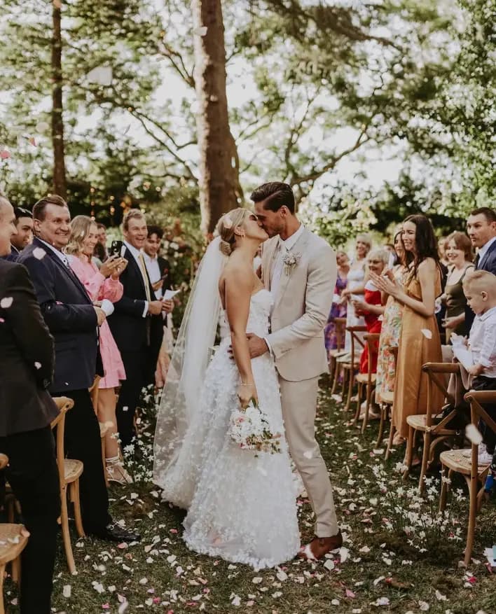 A bride and groom share a kiss at their outdoor wedding ceremony. The bride is wearing a white, intricately designed gown and the groom is in a light beige suit. Guests are seated on either side, applauding and celebrating the couple. Flower petals are scattered on the ground.