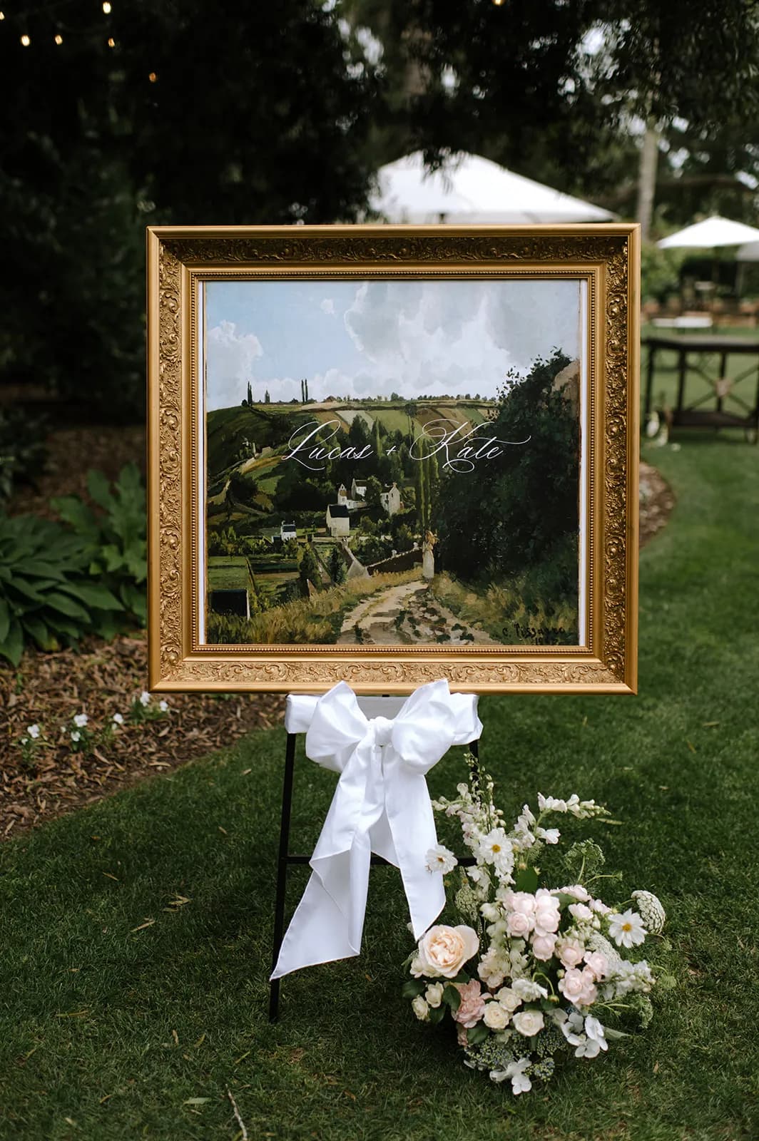 A gold-framed landscape painting with "Luca & Kate" inscribed in elegant white script is displayed on a black easel adorned with a white bow. A bouquet of white and blush flowers lies at the base. The scene is set outdoors with greenery and string lights in the background.