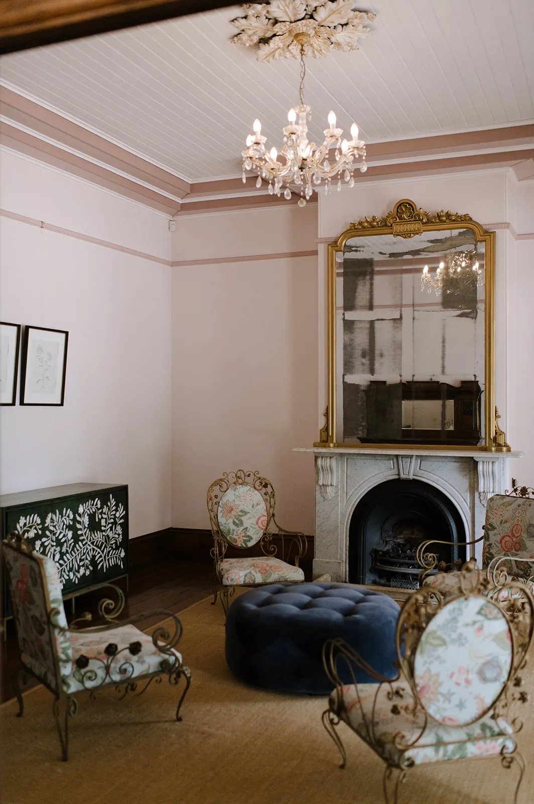 An elegant room features a white ceiling with a chandelier, light pink walls, a large framed mirror above a white fireplace, and four ornate chairs with floral cushions arranged around a blue ottoman. Framed art and a green sideboard with white patterns adorn the room.