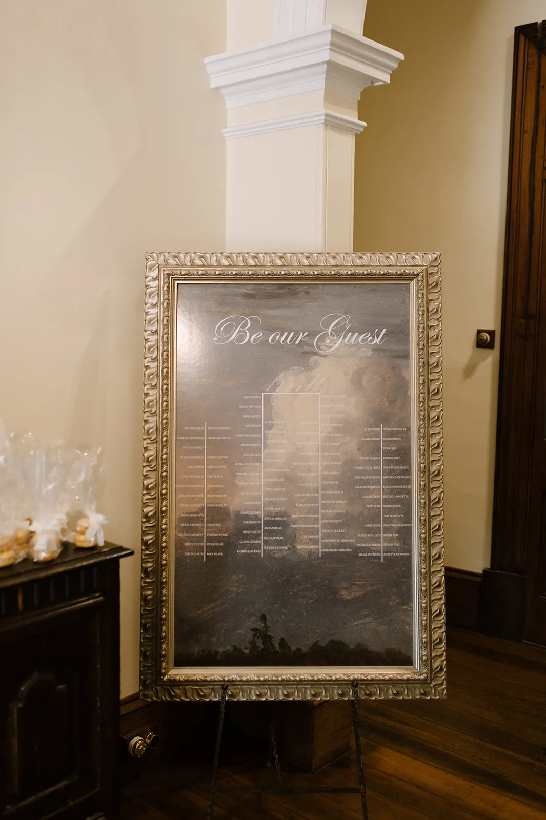 A framed seating chart with "Be our Guest" written at the top, displayed on an easel in an indoor setting. The chart is divided into sections listing names. The background features an artistic, cloudy sky. There's a table with wrapped items to the left.