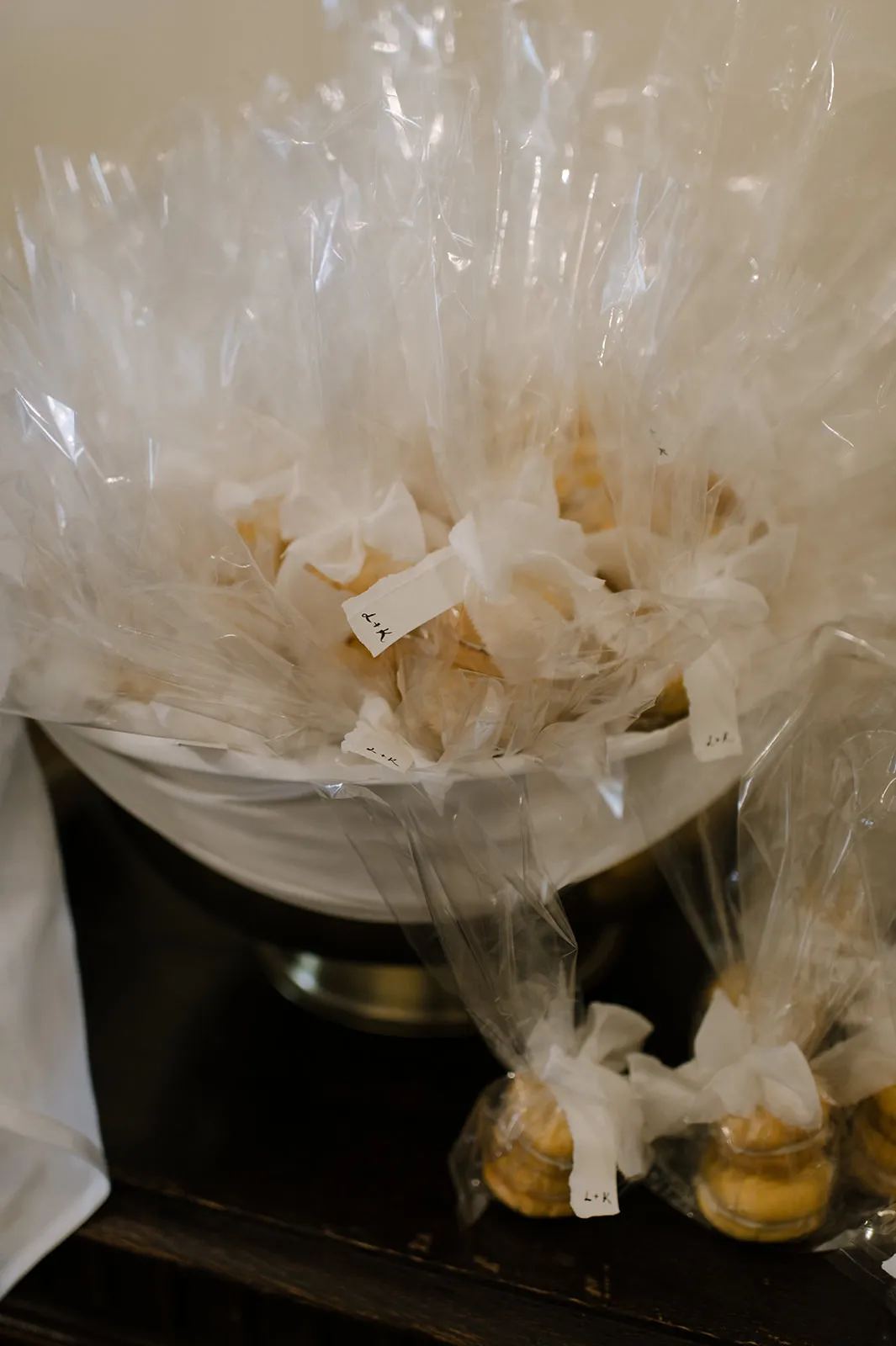 A large white bowl filled with individually wrapped cookies in clear cellophane bags, each tied with white ribbon. The wrapped cookies are arranged to overflow from the bowl, with additional wrapped cookies placed in smaller groups below.