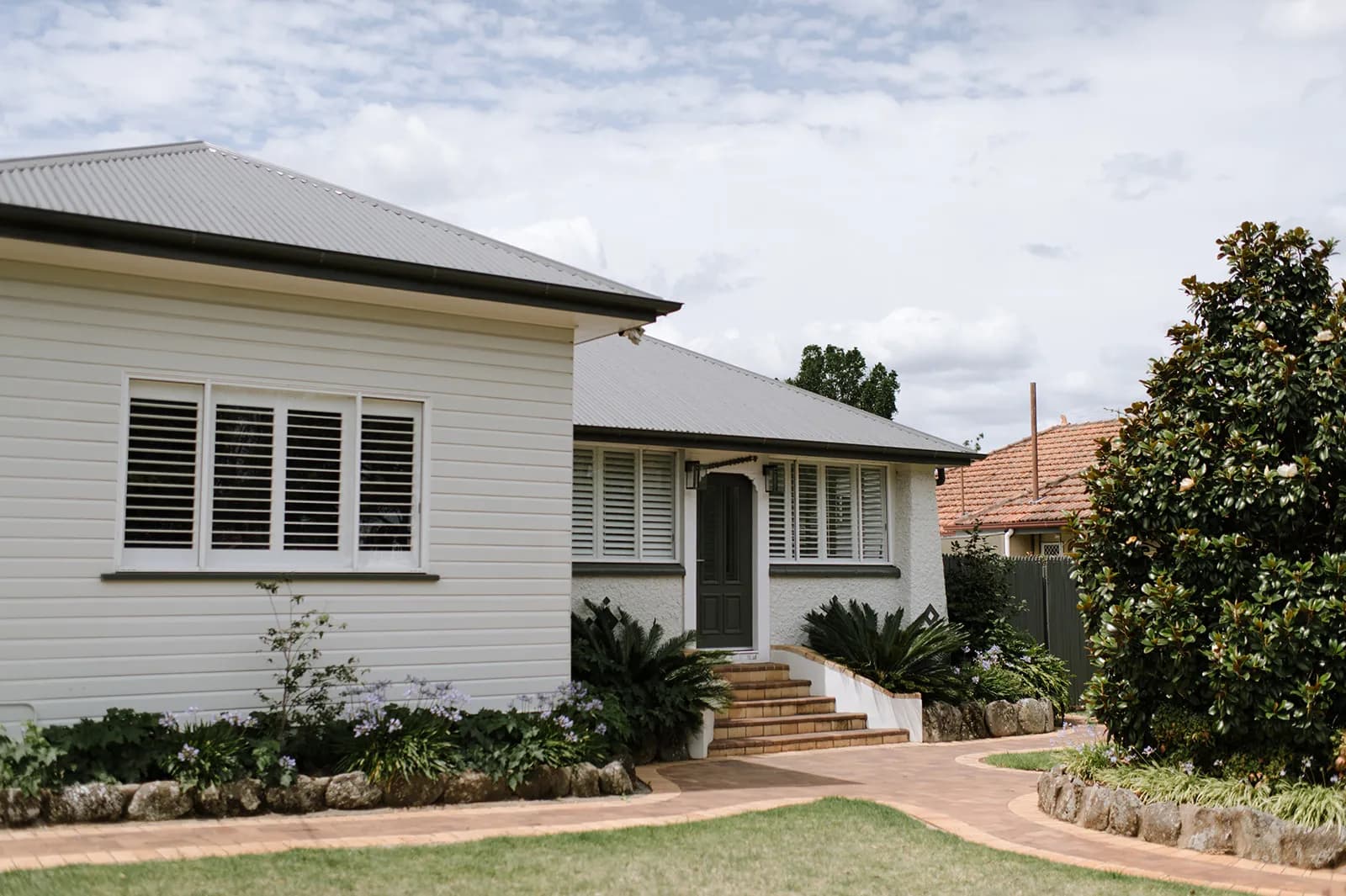 A white single-story house with grey roofing and shutters, surrounded by a well-maintained garden. A brick pathway leads to the entrance, which features a grey door with steps. Trees and shrubs adorn the front yard, and neighboring houses are visible in the background.