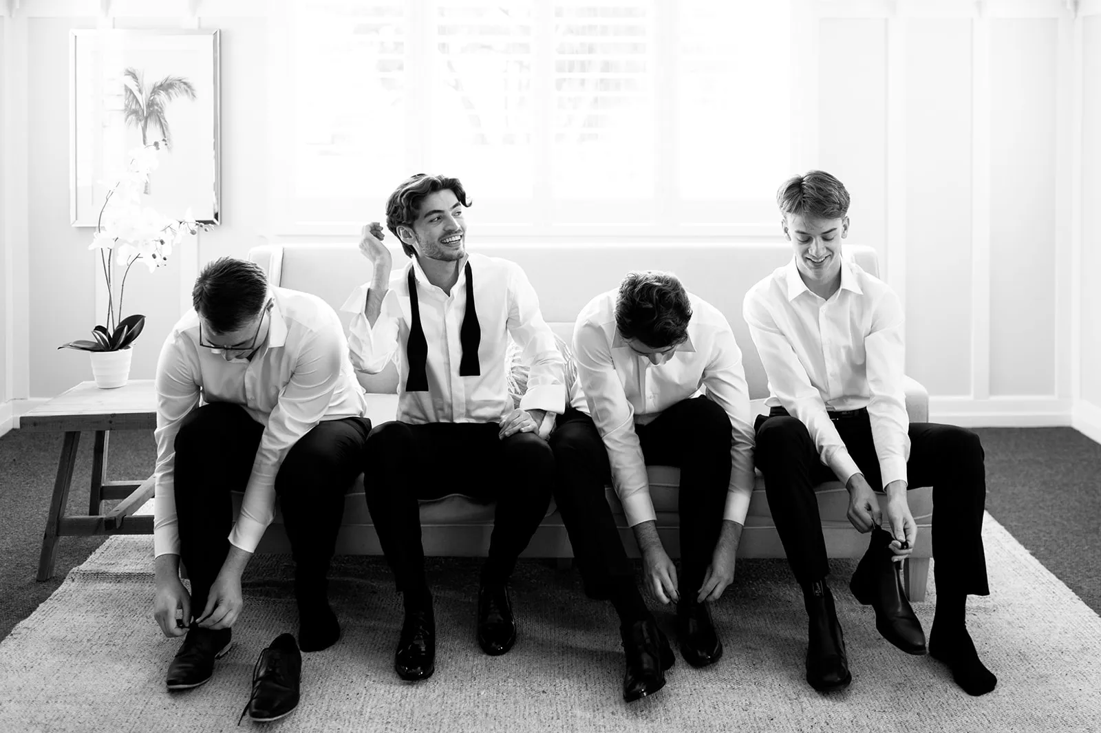 Four men dressed in white shirts are sitting on a couch, preparing for a formal event. Three of them are tying their shoelaces, while one is adjusting his bow tie and smiling. There is a plant in the background near a window, in a bright room.