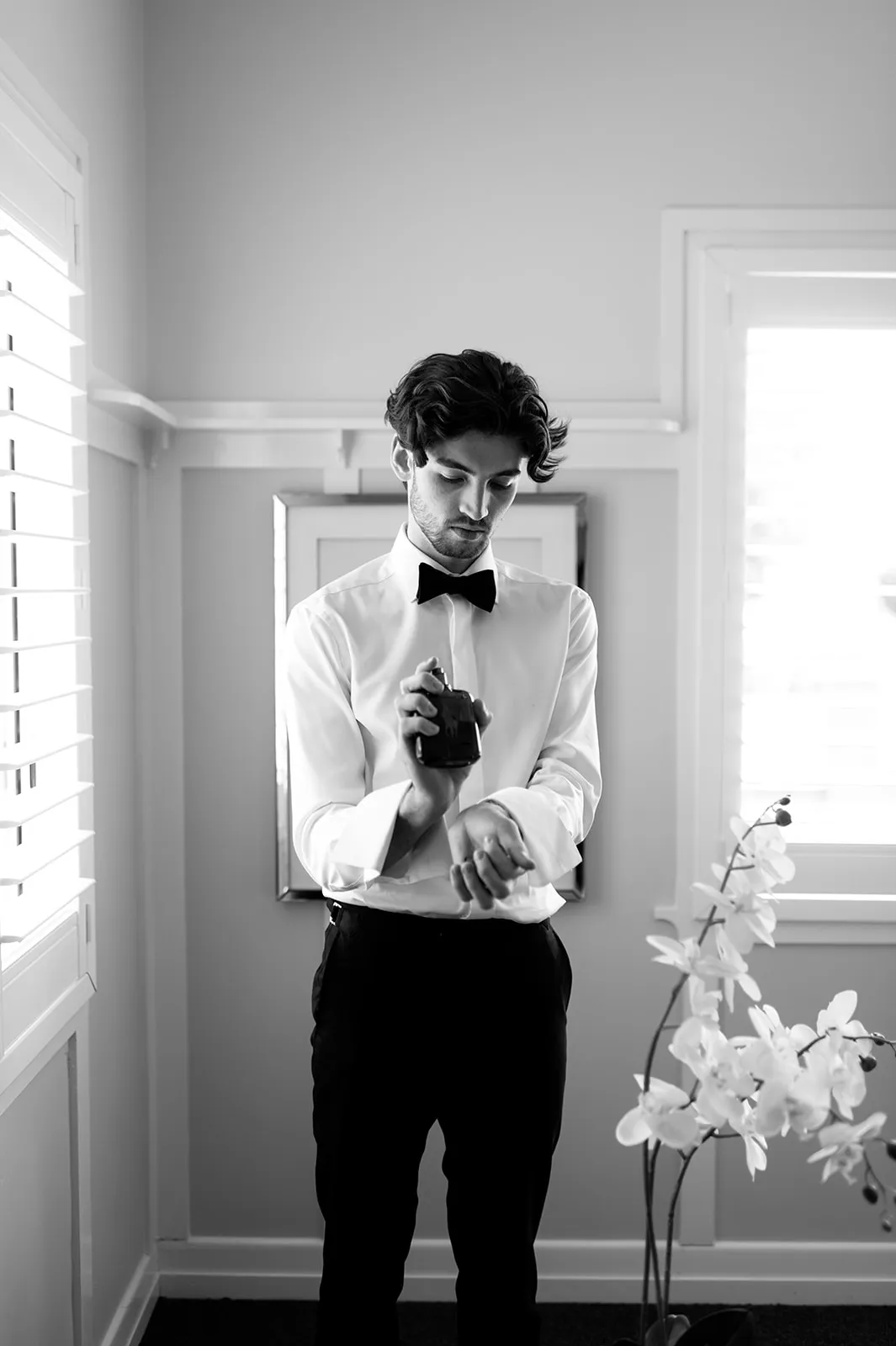 A man dressed in a formal white shirt with a bow tie sprays cologne on his wrist in a bright room. He stands next to a window with shutters and a blooming white orchid, with a mirror hanging on the wall behind him. Black and white image.