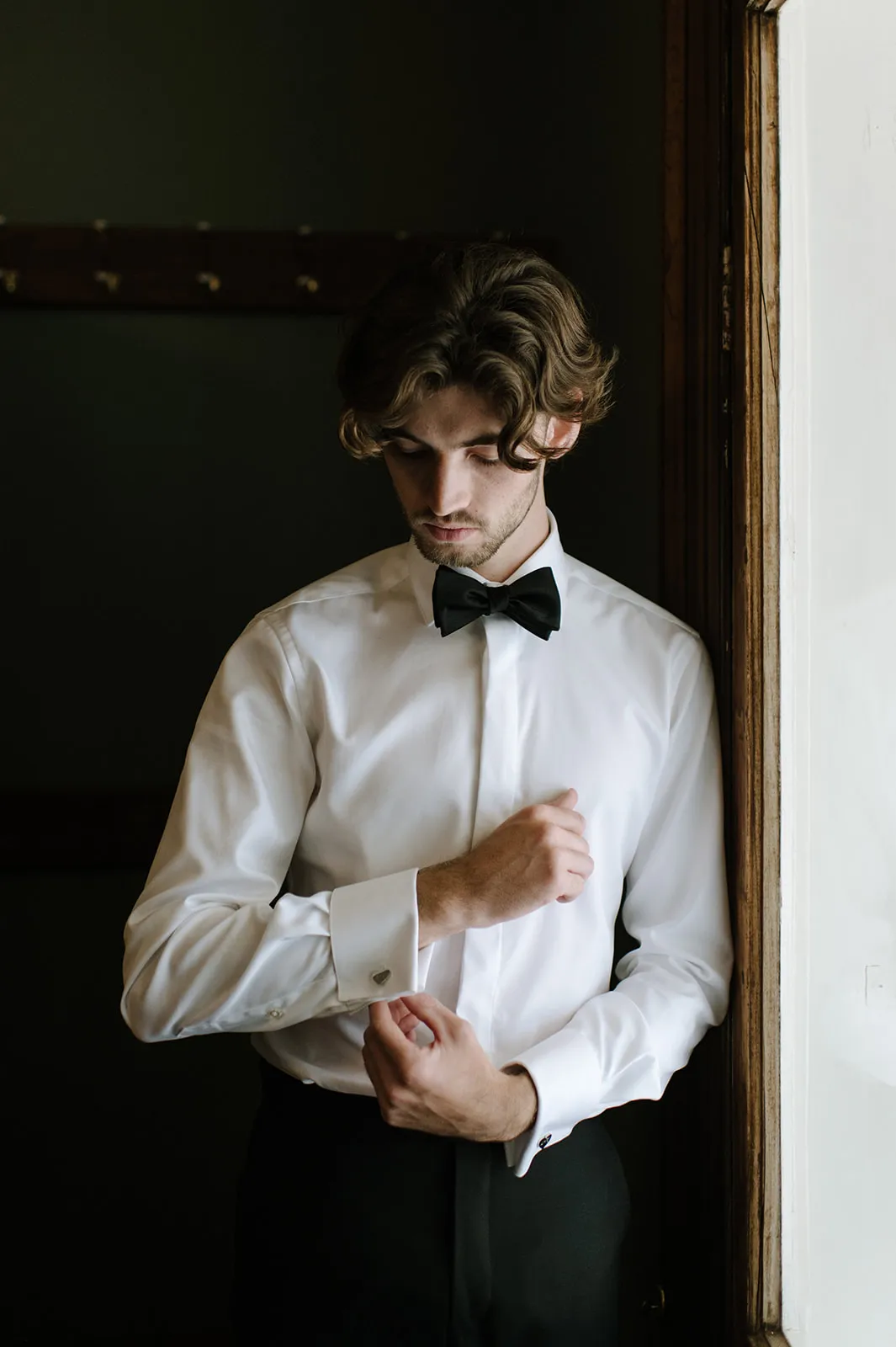 A young man stands by a window adjusting his cufflinks. He is dressed in a white dress shirt, black bow tie, and black pants, looking down with a focused expression. The lighting highlights his attire, casting a soft shadow on his face.