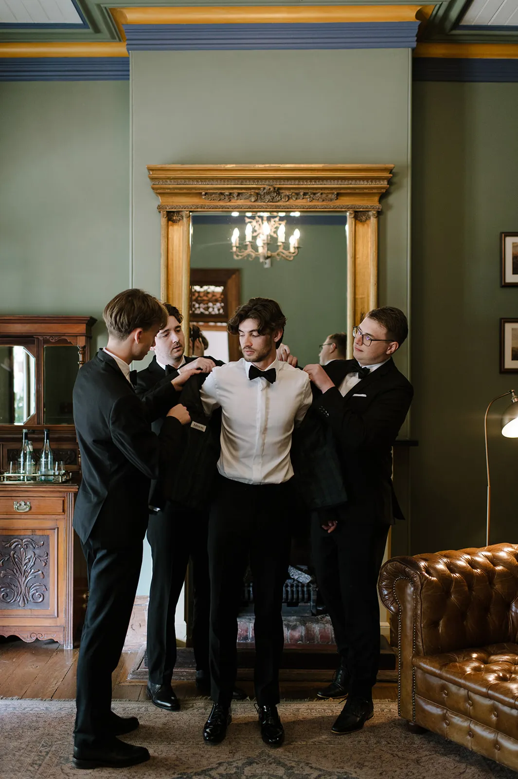 A groom stands in front of a large mirror while three groomsmen help him put on his suit jacket. They are all dressed in formal black suits with bow ties. The room is elegantly decorated with classic wooden furniture, a chandelier, and a leather armchair.