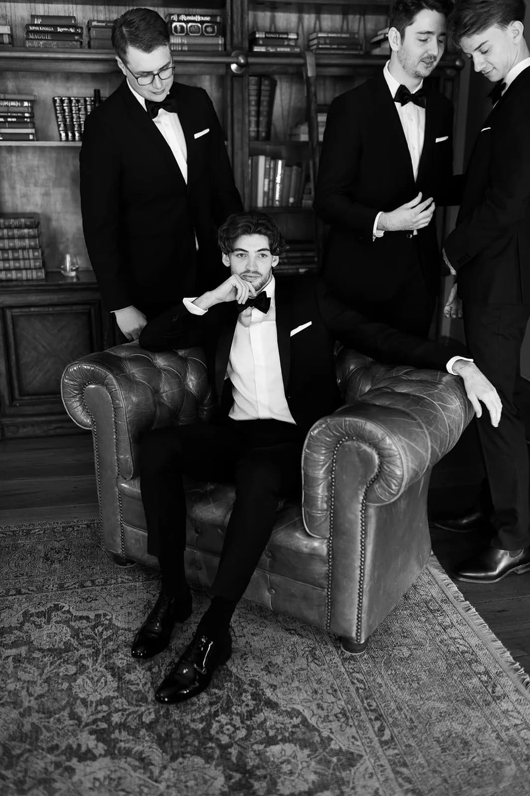 A black-and-white photo features four men dressed in tuxedos. One man is seated in an armchair, looking directly at the camera with a serious expression. The other three men stand behind him, engaged in conversation. They are in a room with shelves filled with books.