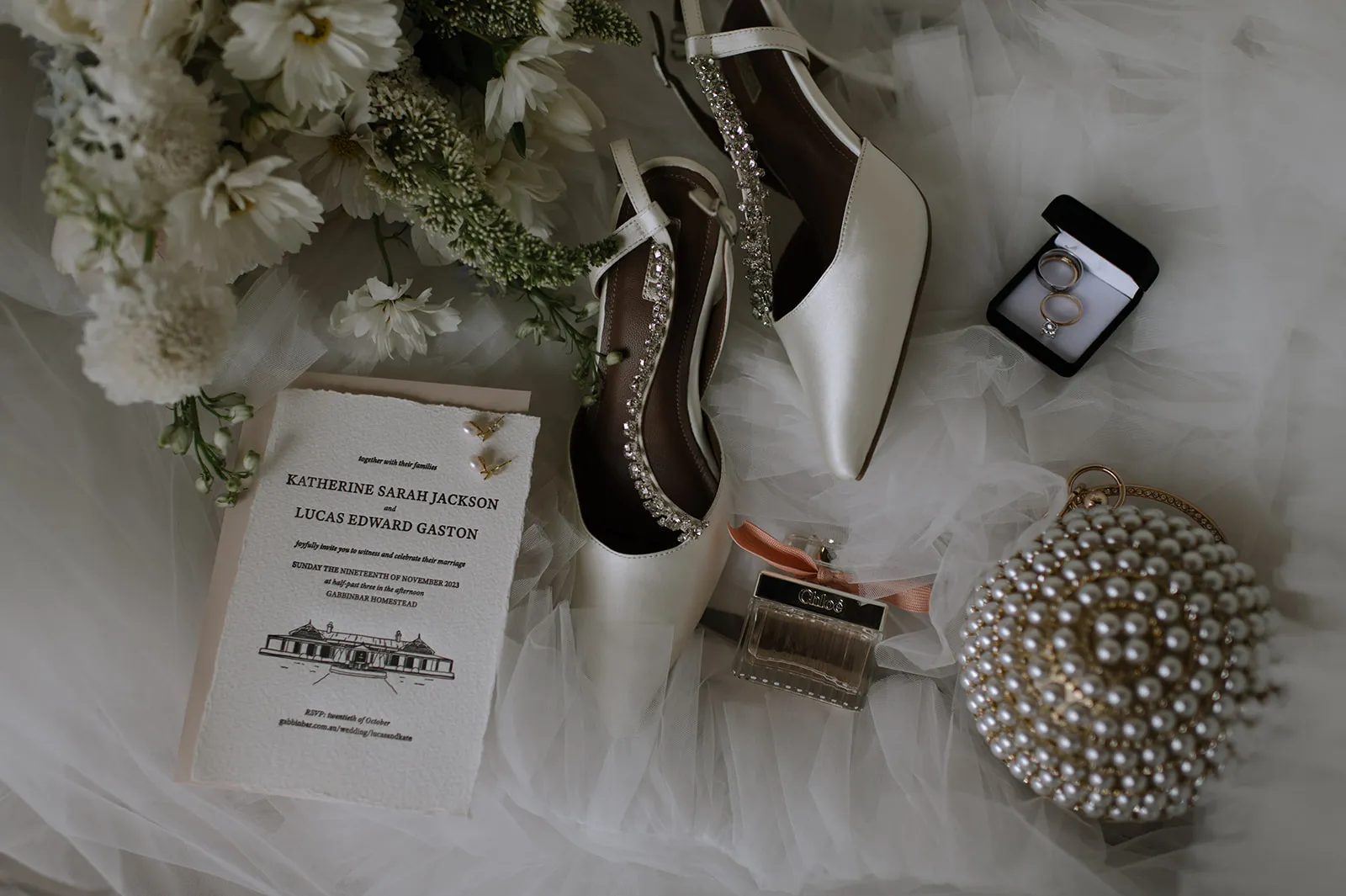 Elegant wedding flat lay featuring a pair of white bridal shoes with jewels, a set of wedding rings in a black box, a wedding invitation, a perfume bottle, a pearl-studded clutch, and a bouquet of white flowers. Text on the invitation includes names and wedding details.