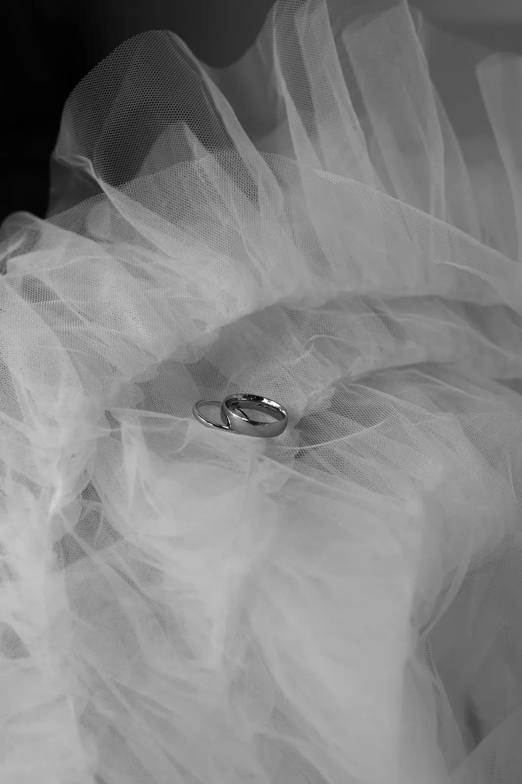 A close-up black and white photograph shows two wedding rings nestled within layers of delicate, translucent tulle fabric. The tulle's soft folds create a dreamy and romantic atmosphere around the shiny rings.