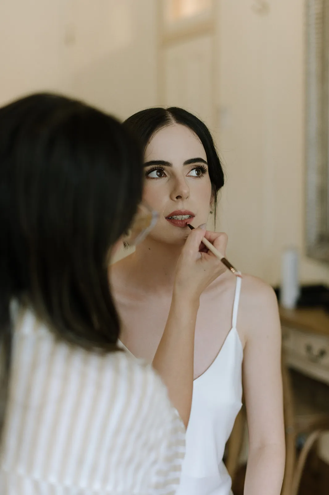 A woman with dark hair in a white dress is having her makeup applied. Another person, partially visible and facing away from the camera, is using a makeup brush to touch up her lips. The scene appears to be indoors in a softly lit room.