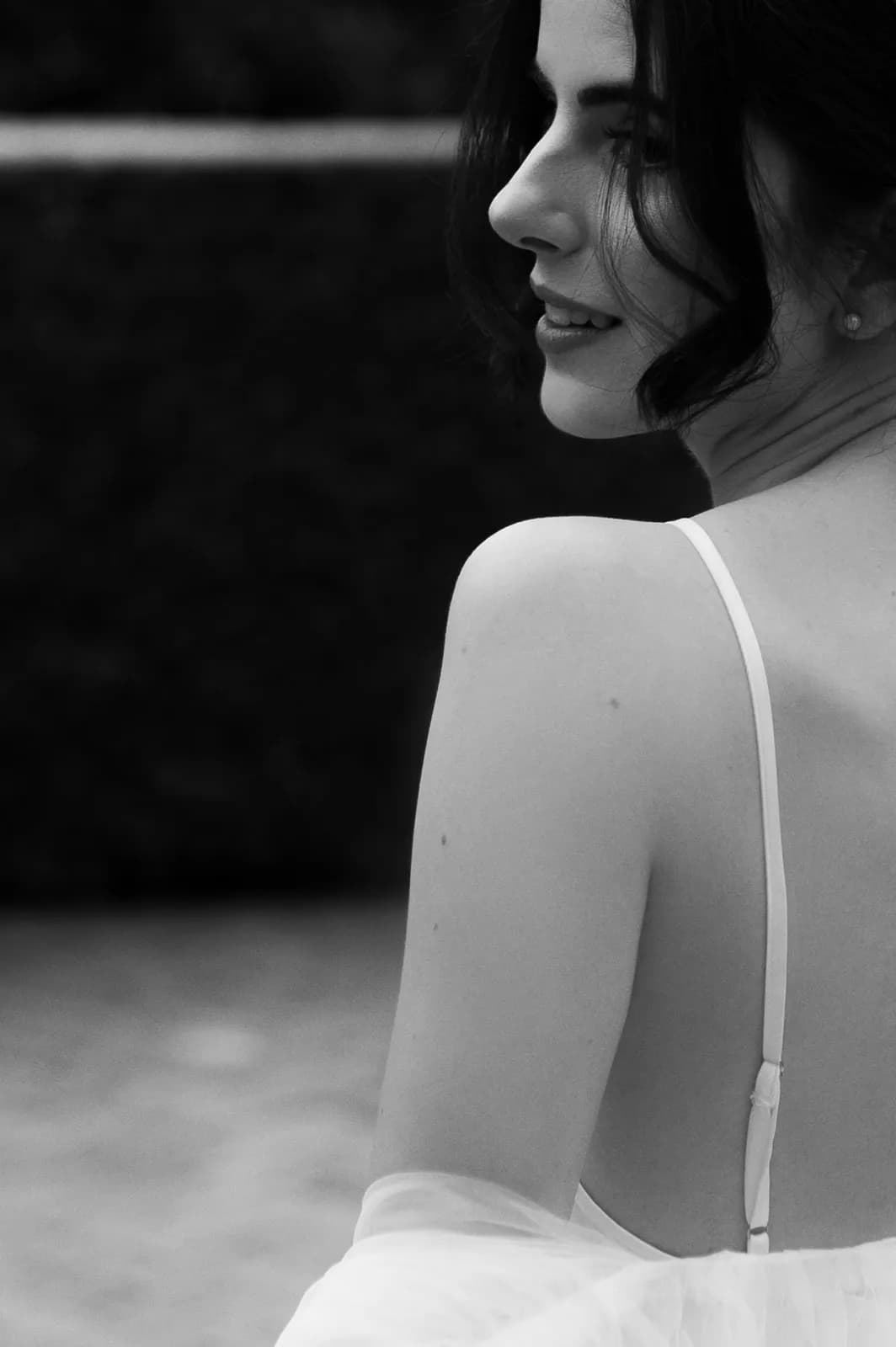 A black-and-white image of a woman with dark hair, looking over her shoulder. She is wearing a thin-strapped top and has a slight smile on her face. The background is blurred, highlighting her sharp profile and elegant posture.