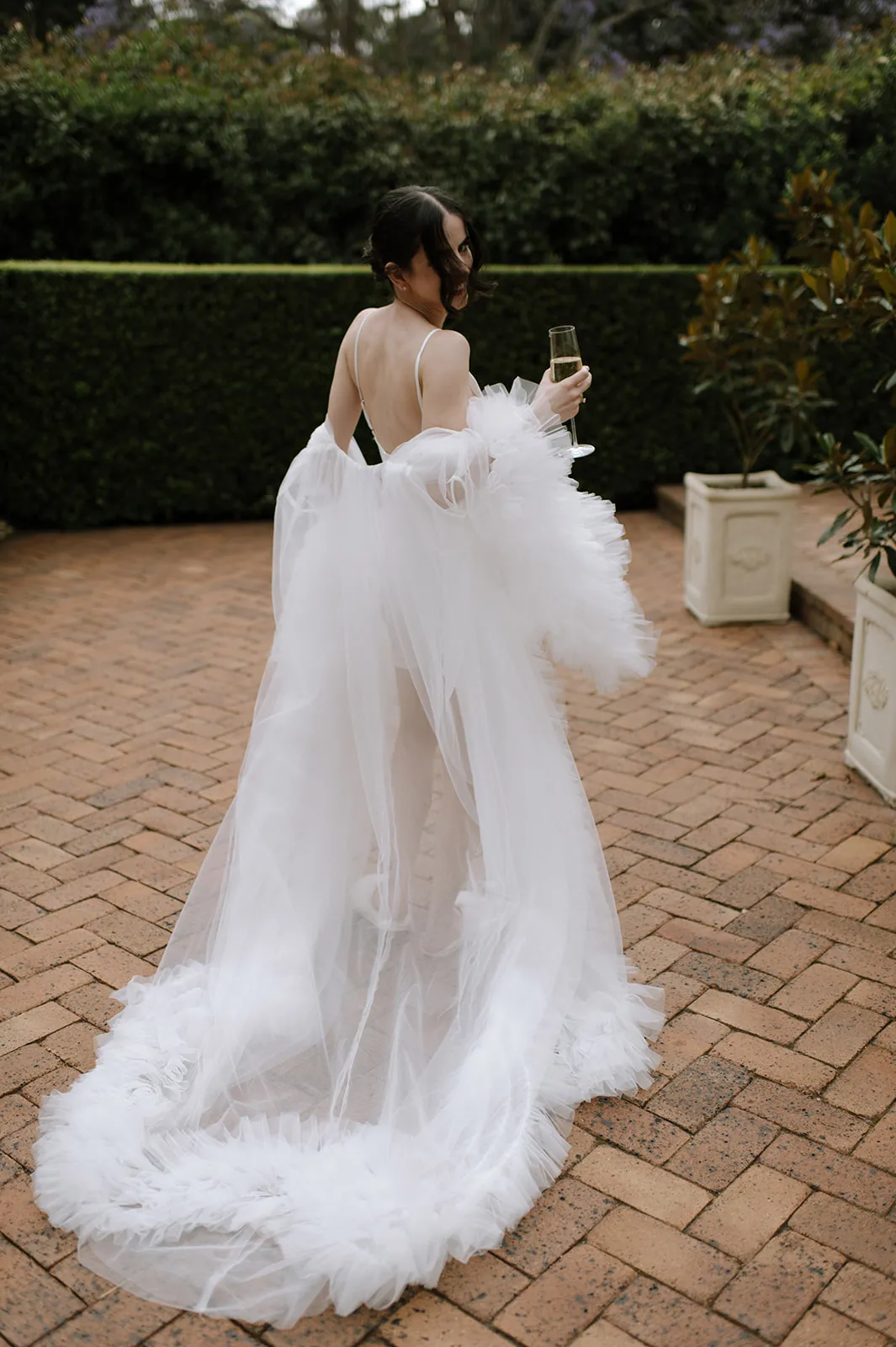 A woman, photographed from behind, stands on a brick pathway surrounded by greenery, holding a flute of champagne. She is wearing an elaborate, flowing white dress with sheer fabric and ruffled details, giving a soft and ethereal appearance.