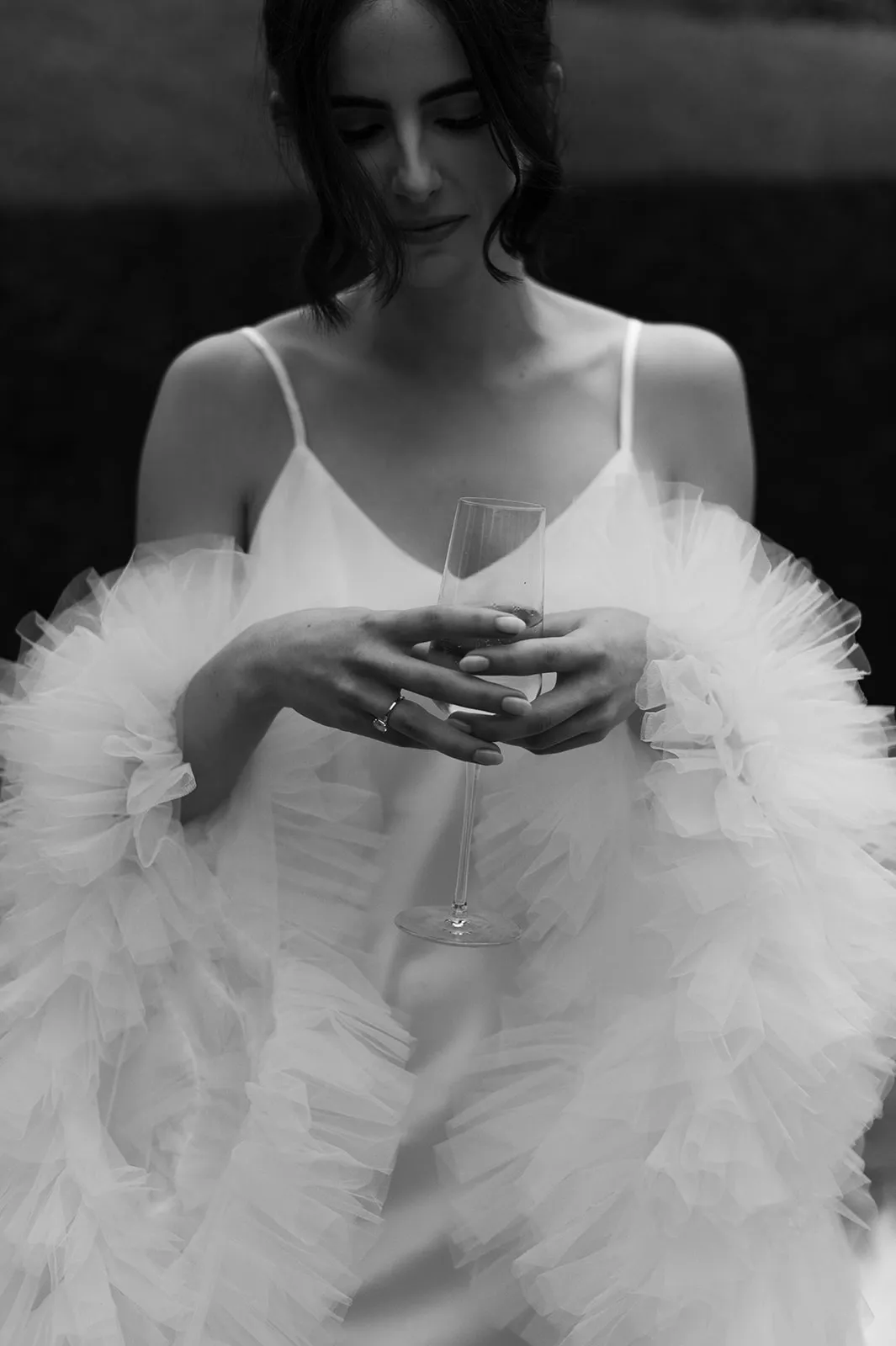 Black-and-white photo of a woman wearing a sleeveless, ruffled dress, holding a champagne glass between her fingers. Her gaze is downcast, and she has shoulder-length hair styled in loose waves. The background is dark and out of focus, emphasizing her ethereal appearance.