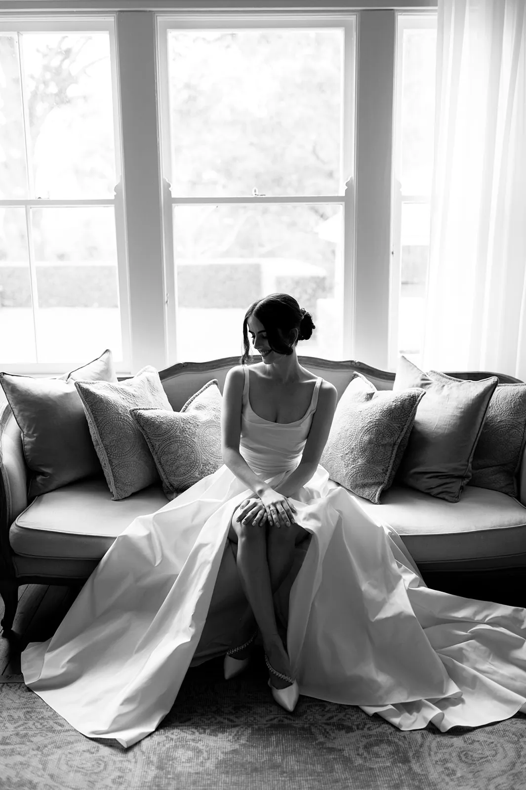 A black and white photo of a bride in a flowing wedding gown sitting on a couch. She is looking down, hands clasped, with her long dress spread out around her. The couch is adorned with several pillows and a large window is behind her, letting in natural light.