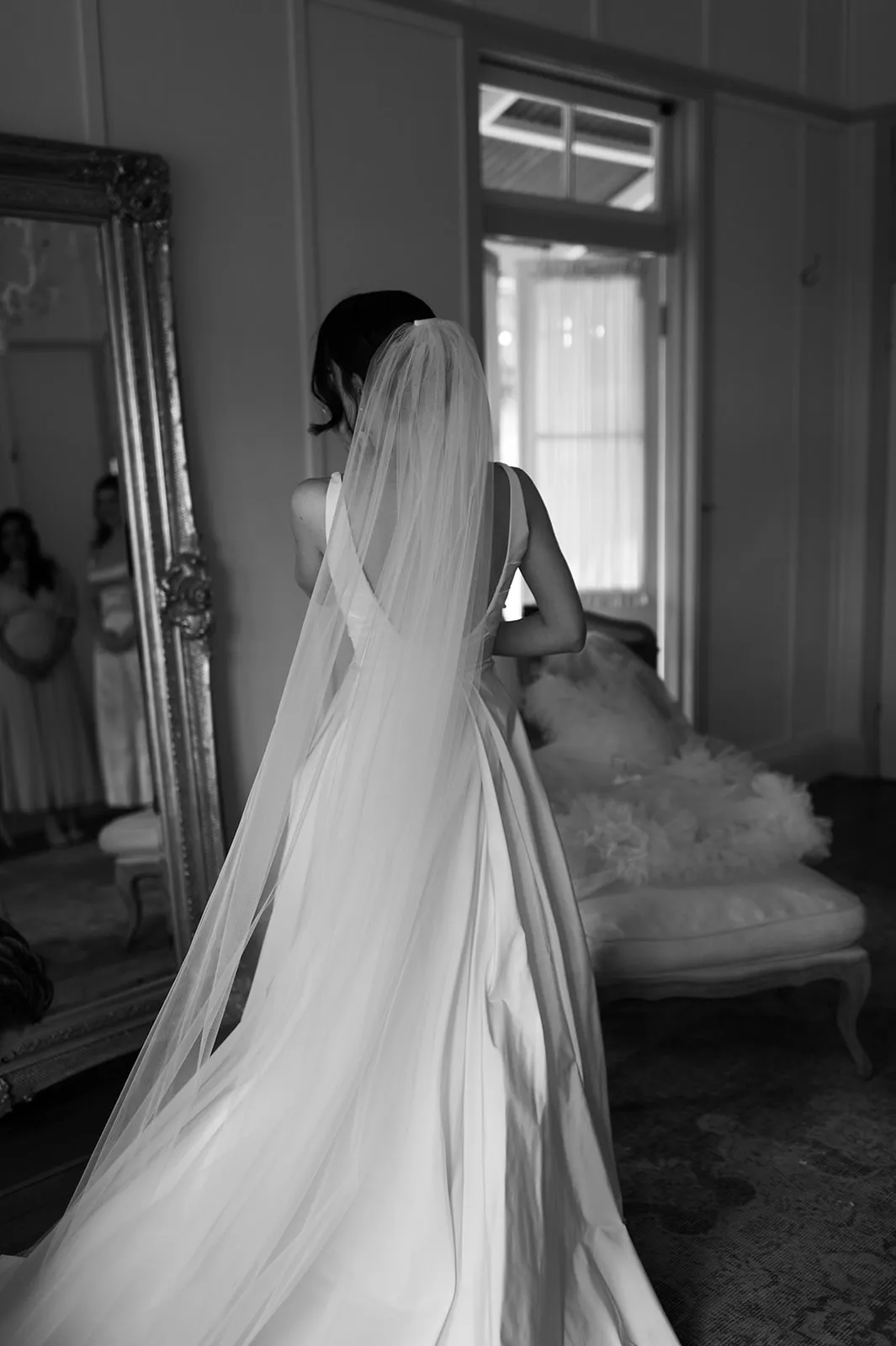 A black and white photo of a bride in a wedding dress with a long veil, seen from the back. She stands in front of a mirror, with two bridesmaids visible in the background. The room has elegant decor with a sofa and draped fabric.