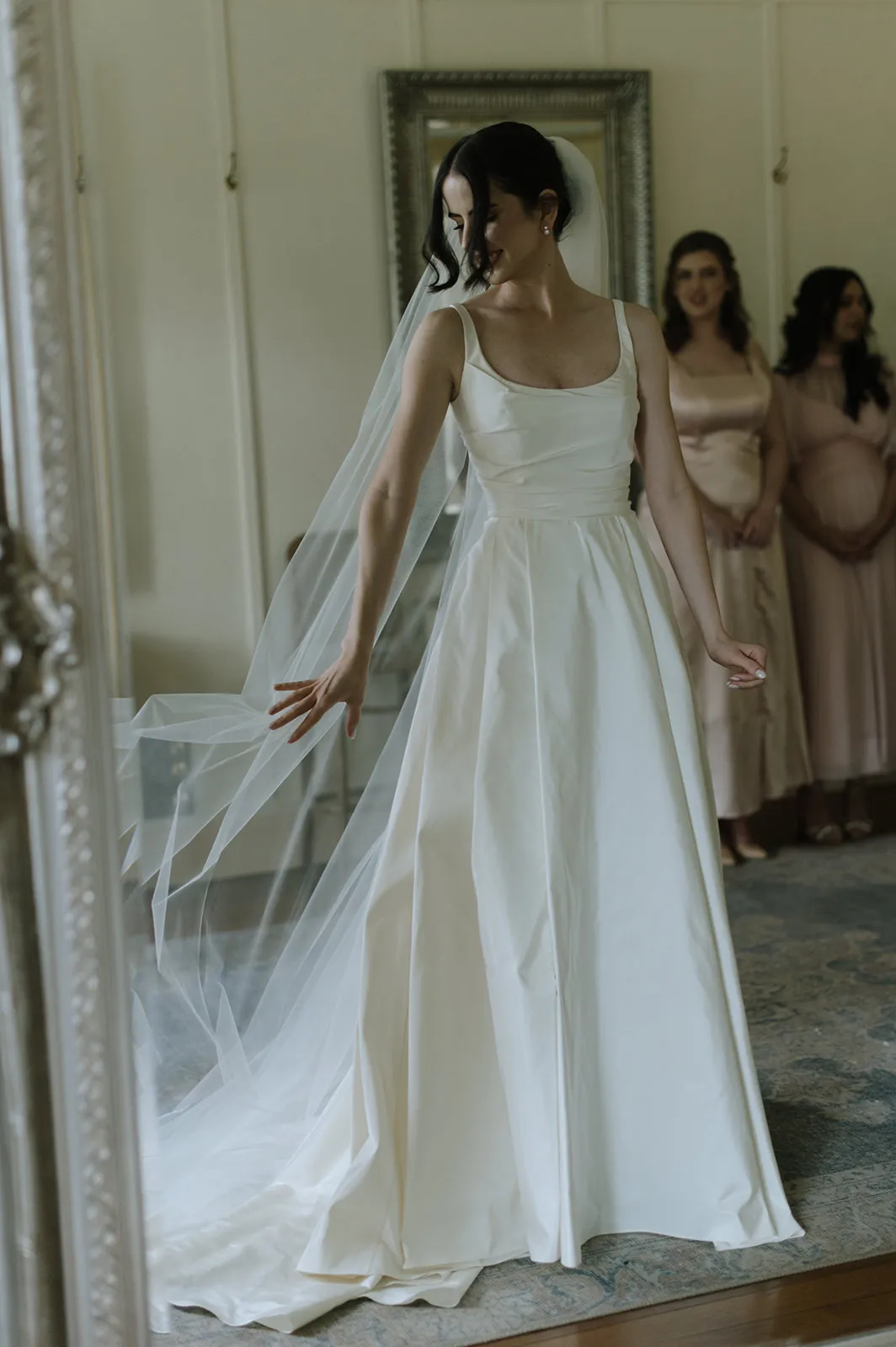 A bride in a white gown and long veil stands gracefully, looking down and gently touching the fabric of her dress. Behind her, two bridesmaids in blush gowns stand, with one visibly pregnant. The room is softly lit, with a large mirror reflecting part of the scene.