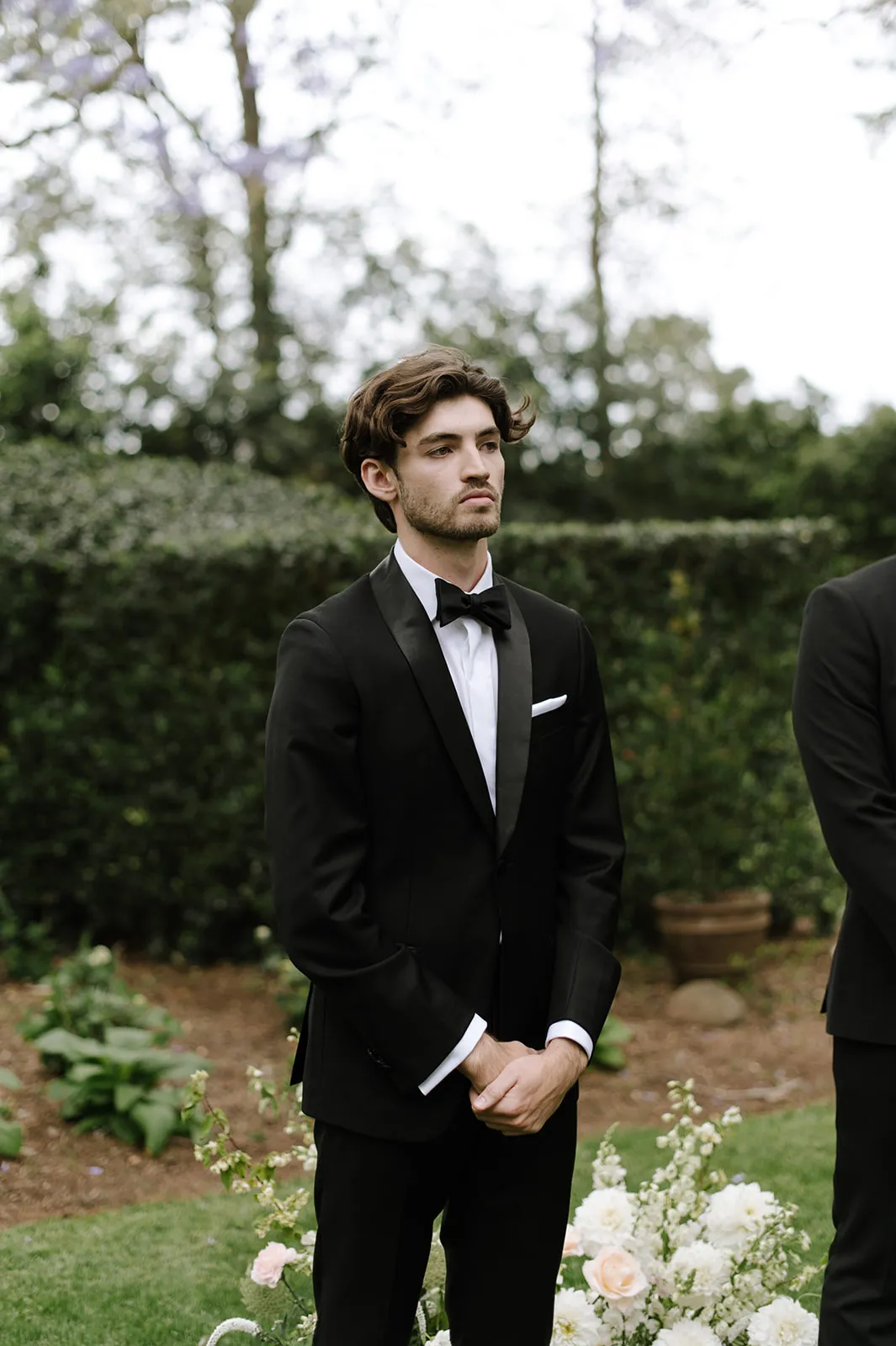 A man dressed in a black tuxedo with a white pocket square and a black bow tie stands with his hands folded in front of him in an outdoor setting. The background includes lush greenery and some floral arrangements. He has a serious expression on his face.