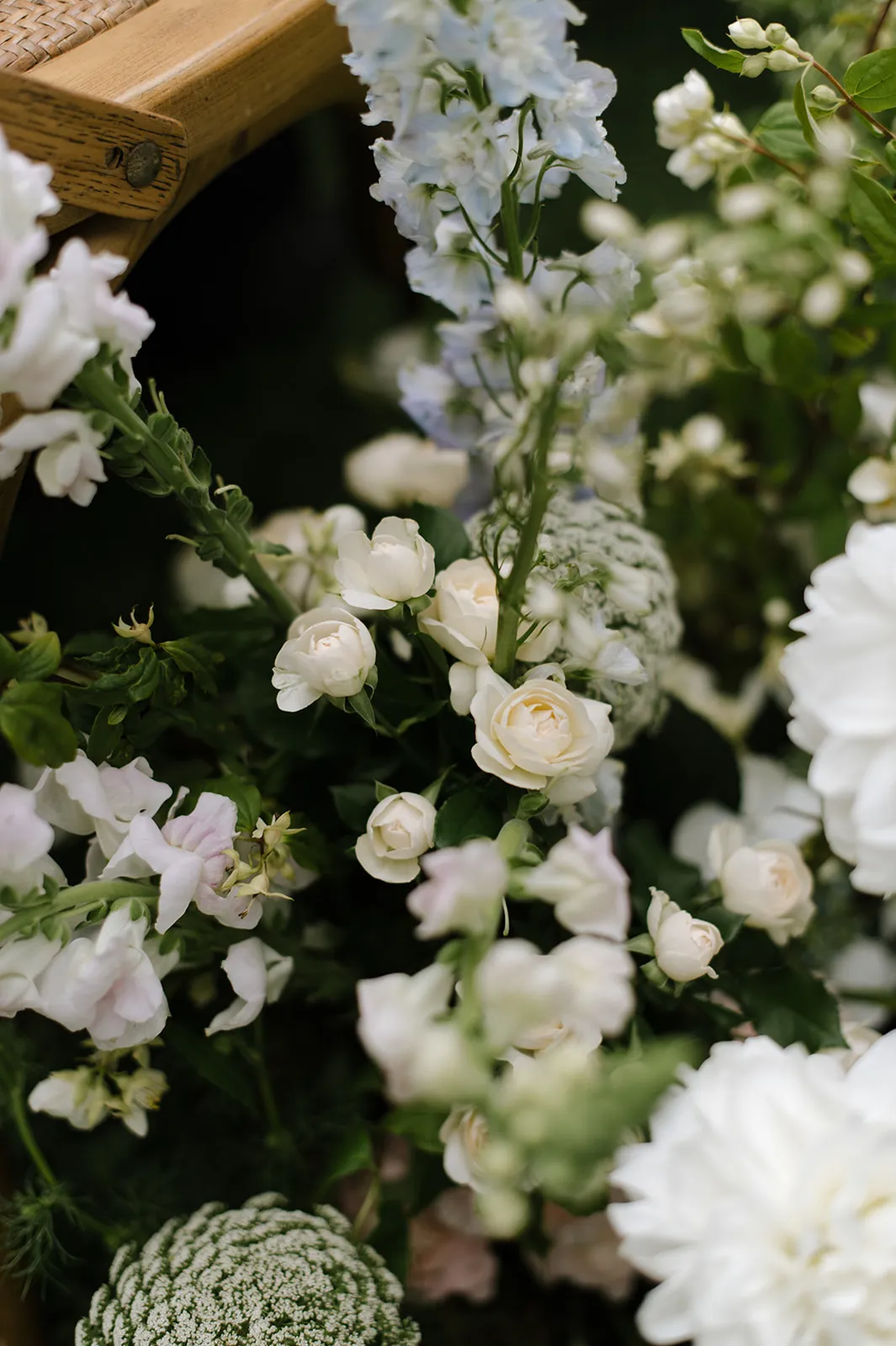 A close-up of a variety of flowers including white roses, delicate white and pink blooms, and pale blue flowers. The flowers are arranged naturally with green foliage, creating a soft, lush display. The corner of a wooden surface is visible in the top left of the image.