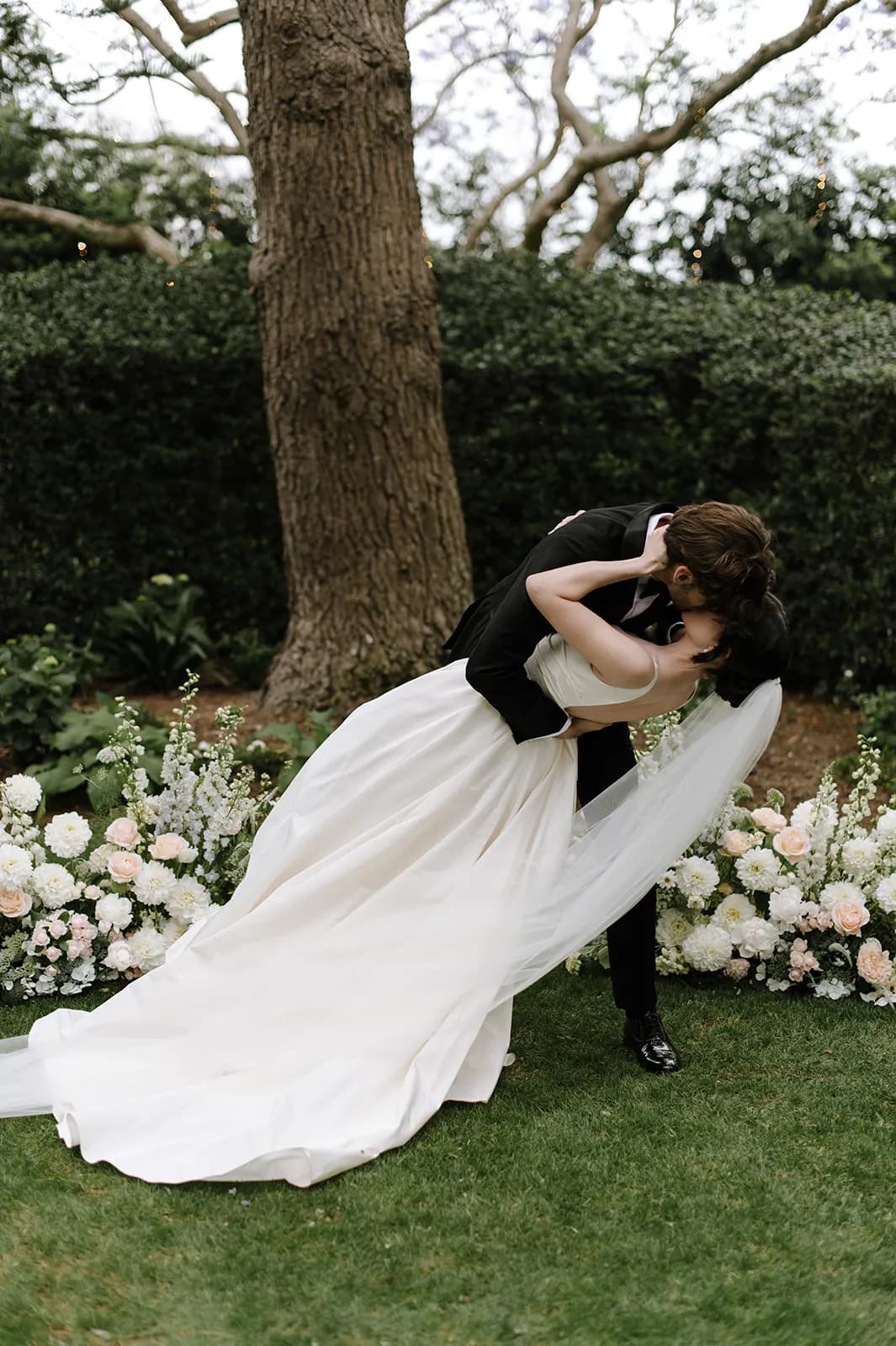 A newlywed couple shares a kiss outdoors, with the groom dipping the bride backwards. They are surrounded by lush greenery and an array of white and pale pink flowers at their feet. The bride's flowing dress and veil contrast with the groom's black suit.