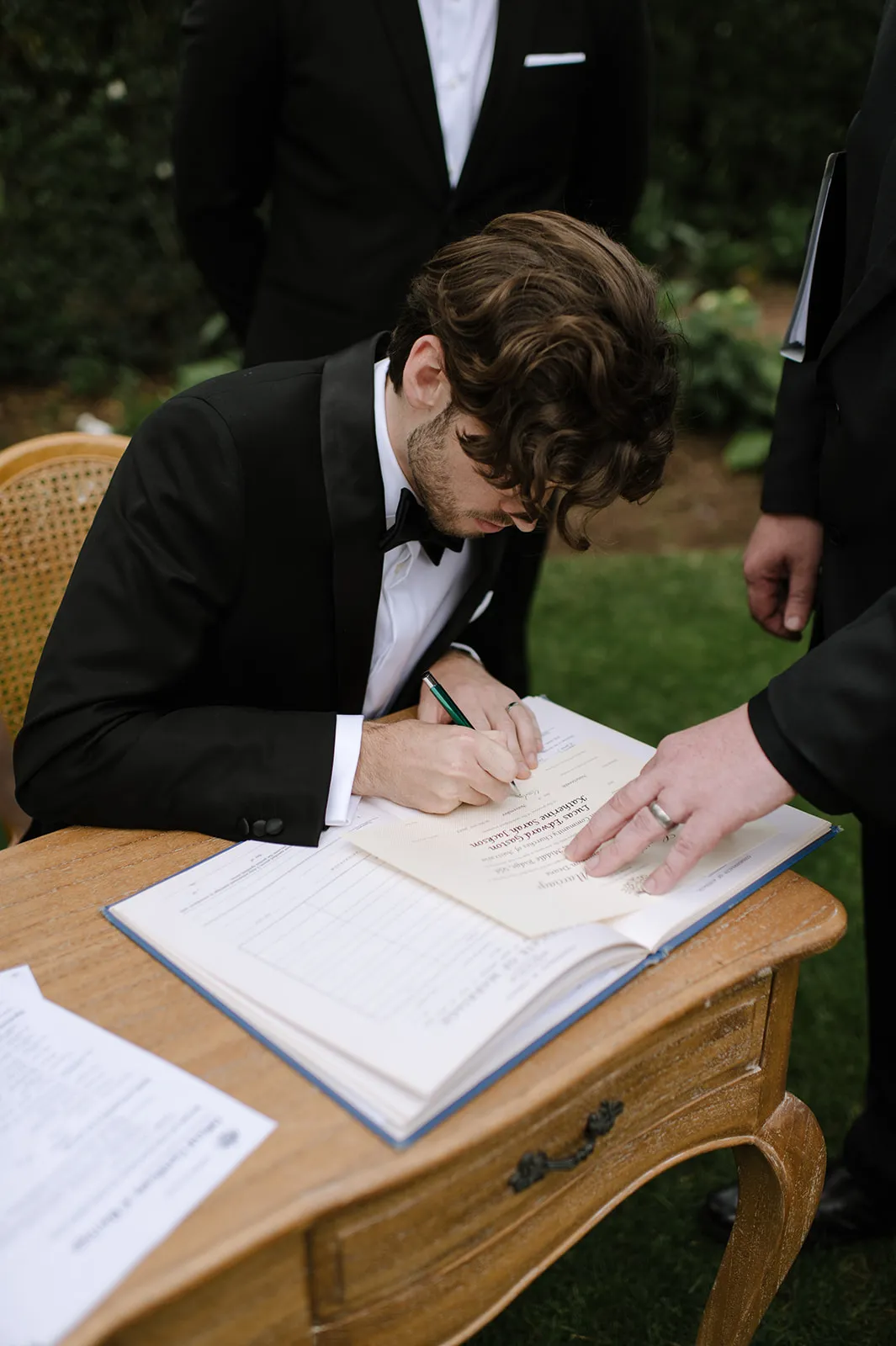 A man in a black tuxedo with wavy brown hair is bending over a wooden table, signing a large book. The scene is set outdoors on a grassy area. Another person’s hand and torso are visible next to him, also in a black suit.