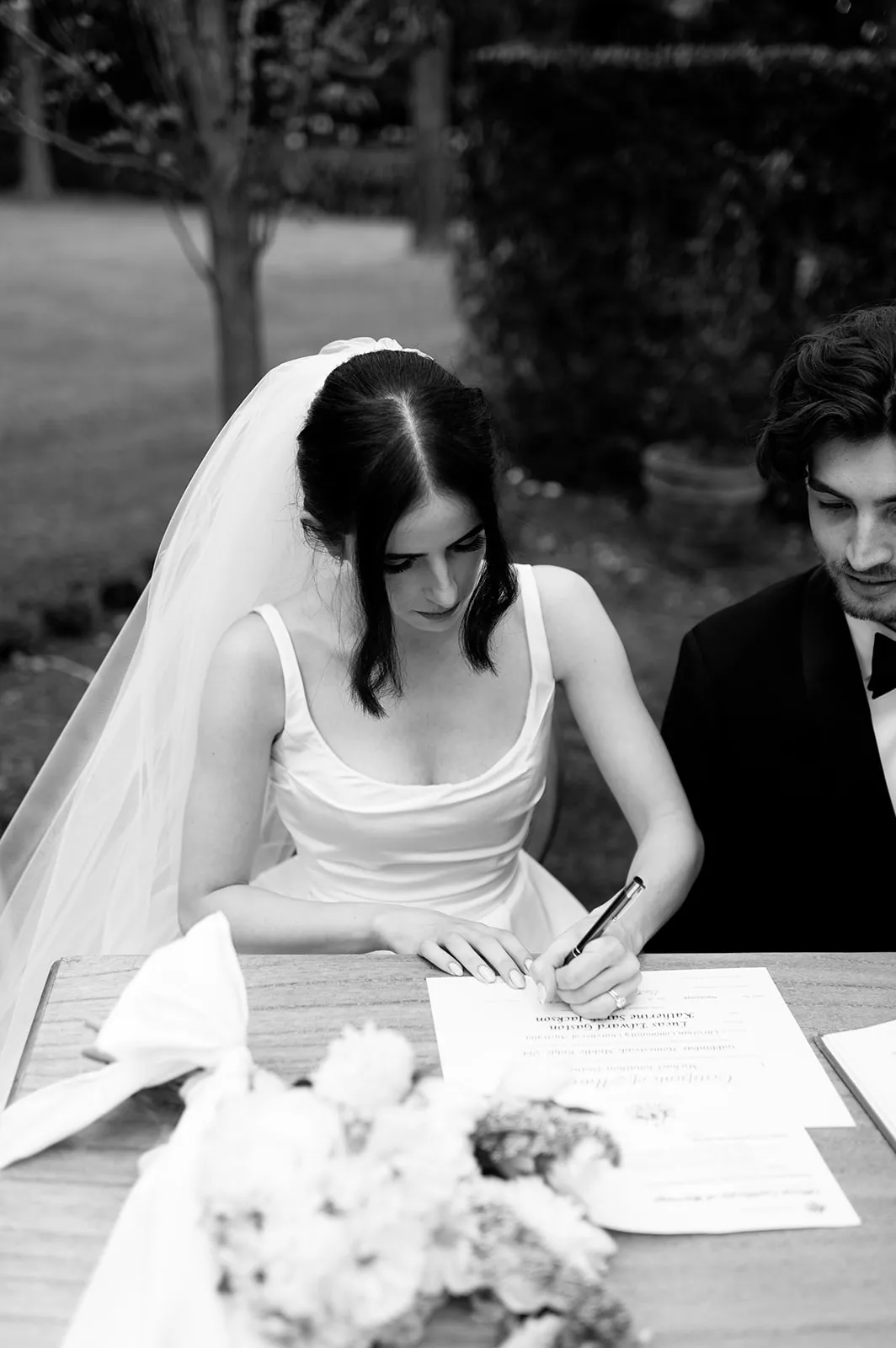 A bride wearing a white wedding dress and veil signs a document at a table adorned with flowers while a groom in a suit sits beside her, outdoors.