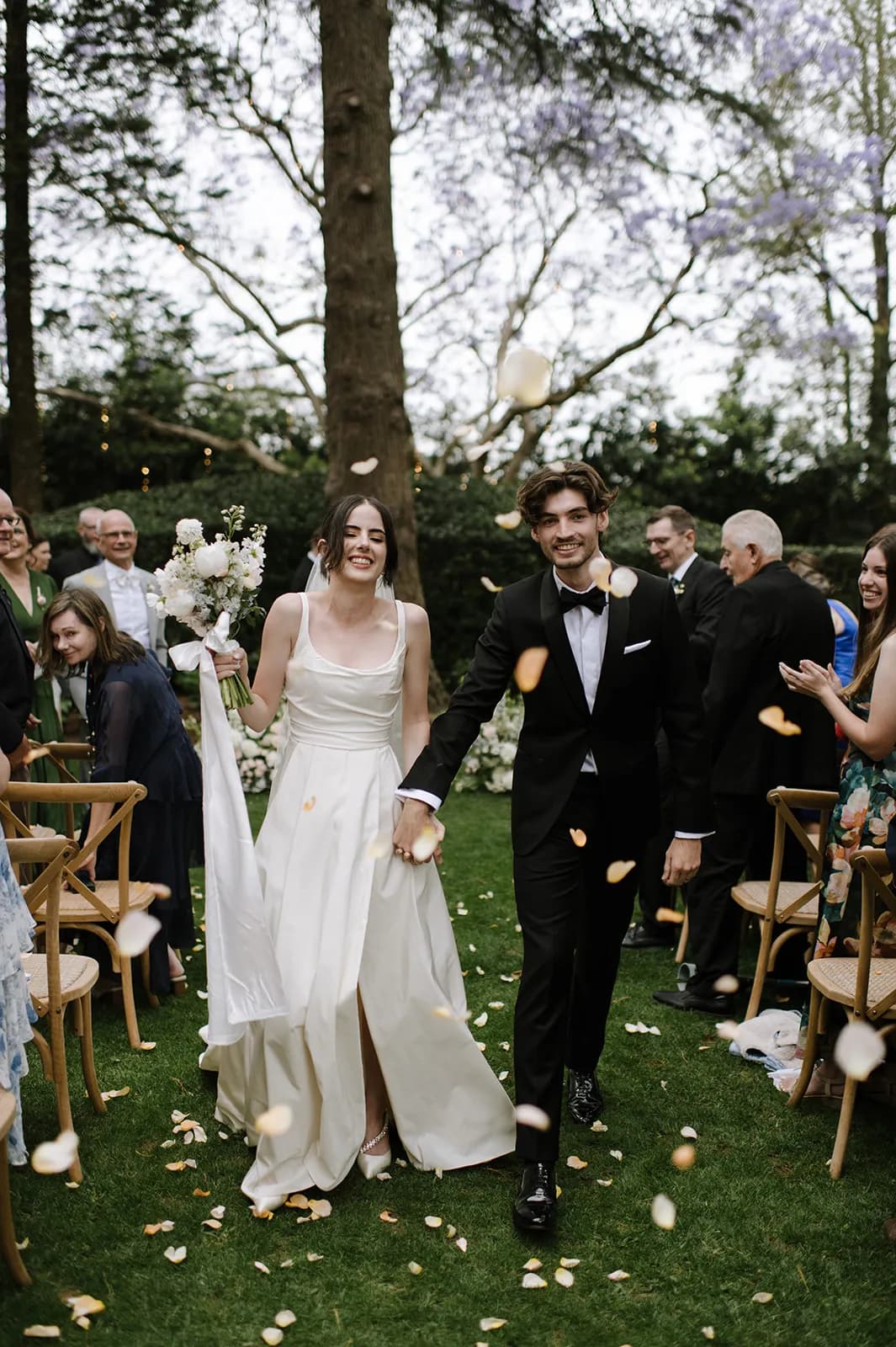 A bride in a white dress and a groom in a black tuxedo walk hand in hand down an outdoor aisle as guests throw flower petals. They are both smiling joyfully. Guests are seated on either side, clapping and celebrating. Trees and string lights are visible in the background.