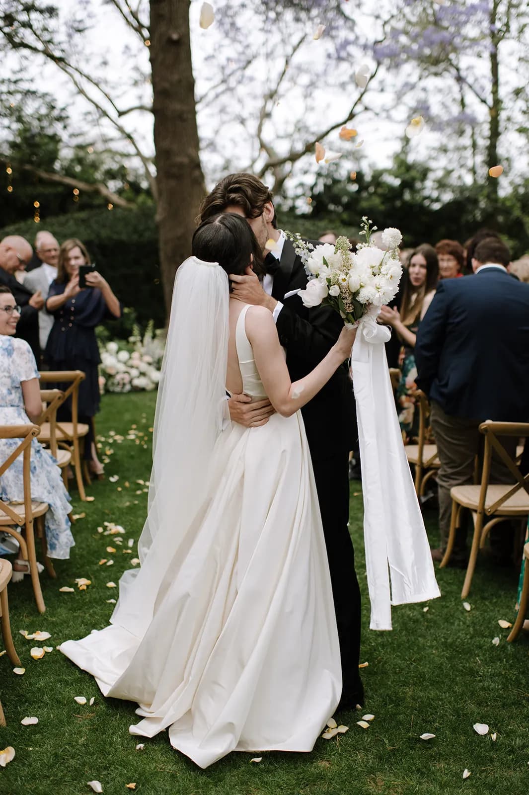 A newlywed couple shares a kiss outdoors in a garden setting with decorated trees in the background. The bride wears a white gown and veil while holding a bouquet of white flowers. Guests are seated and standing around the couple, witnessing the moment.