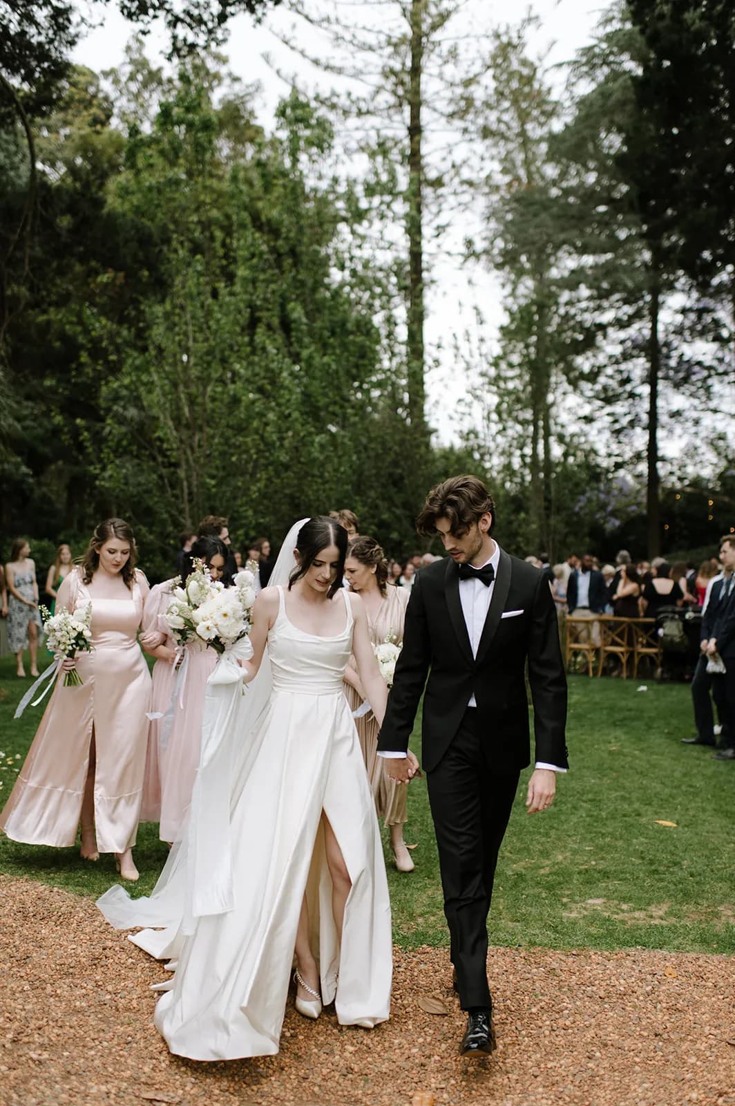 A bride in a white gown and a groom in a black tuxedo walk hand in hand outdoors, followed by bridesmaids in pink dresses holding bouquets. They are surrounded by trees and a gathering of people in the background, all part of a wedding celebration.