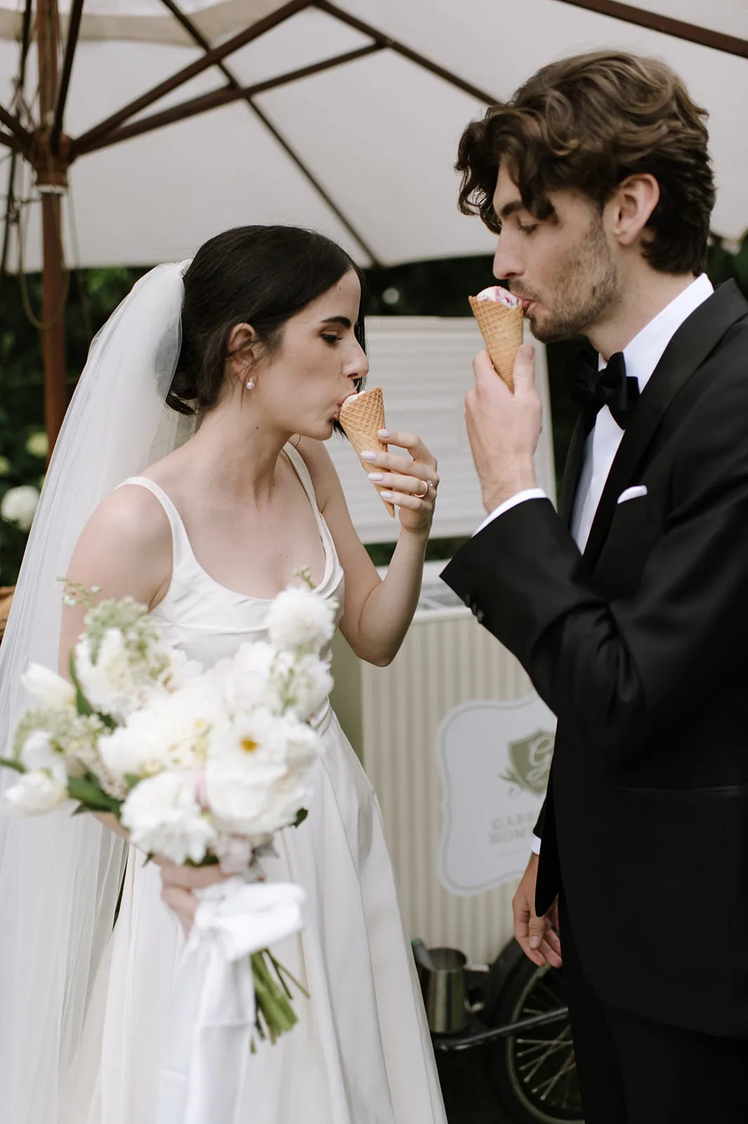 A bride and groom, dressed in wedding attire, enjoy ice cream cones together under a large umbrella. The bride holds a bouquet of flowers, while both take a bite of their ice cream. The scene suggests a casual and joyful moment during their wedding celebration.