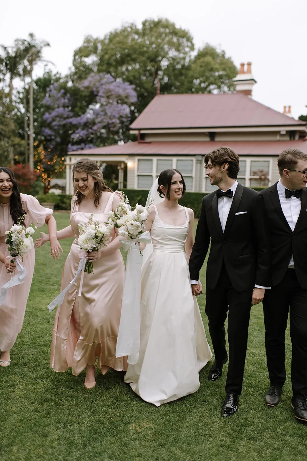 A bride and groom walk hand in hand with their wedding party on a lawn. The bride wears a white gown and the groom is in a black tuxedo. Bridesmaids in pink dresses and groomsmen in black suits walk alongside. Trees and a house are visible in the background.